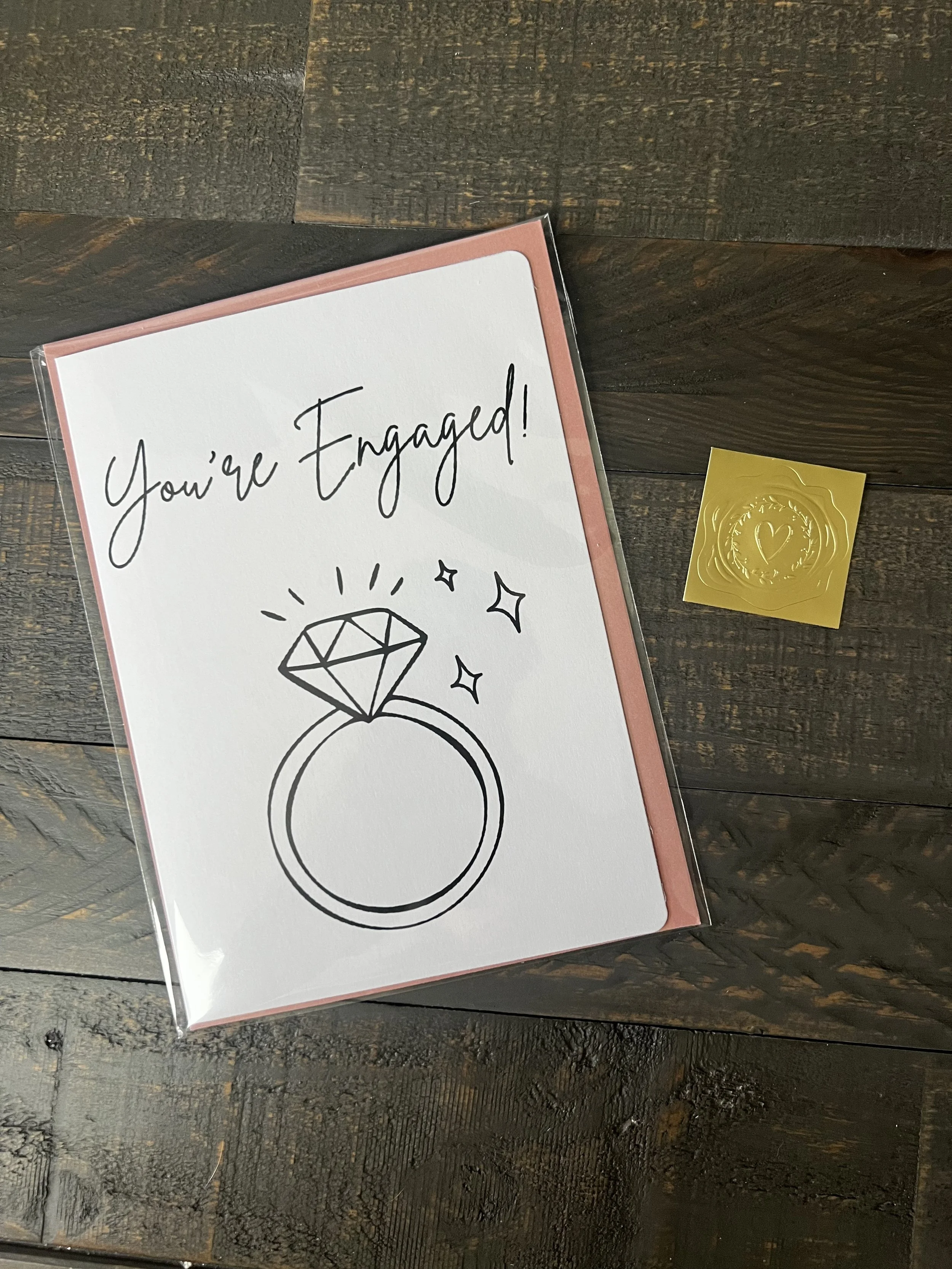 Styled flat lay of You're Engaged! greeting card with coordinating blush pink envelope and gold wax seal style sticker.