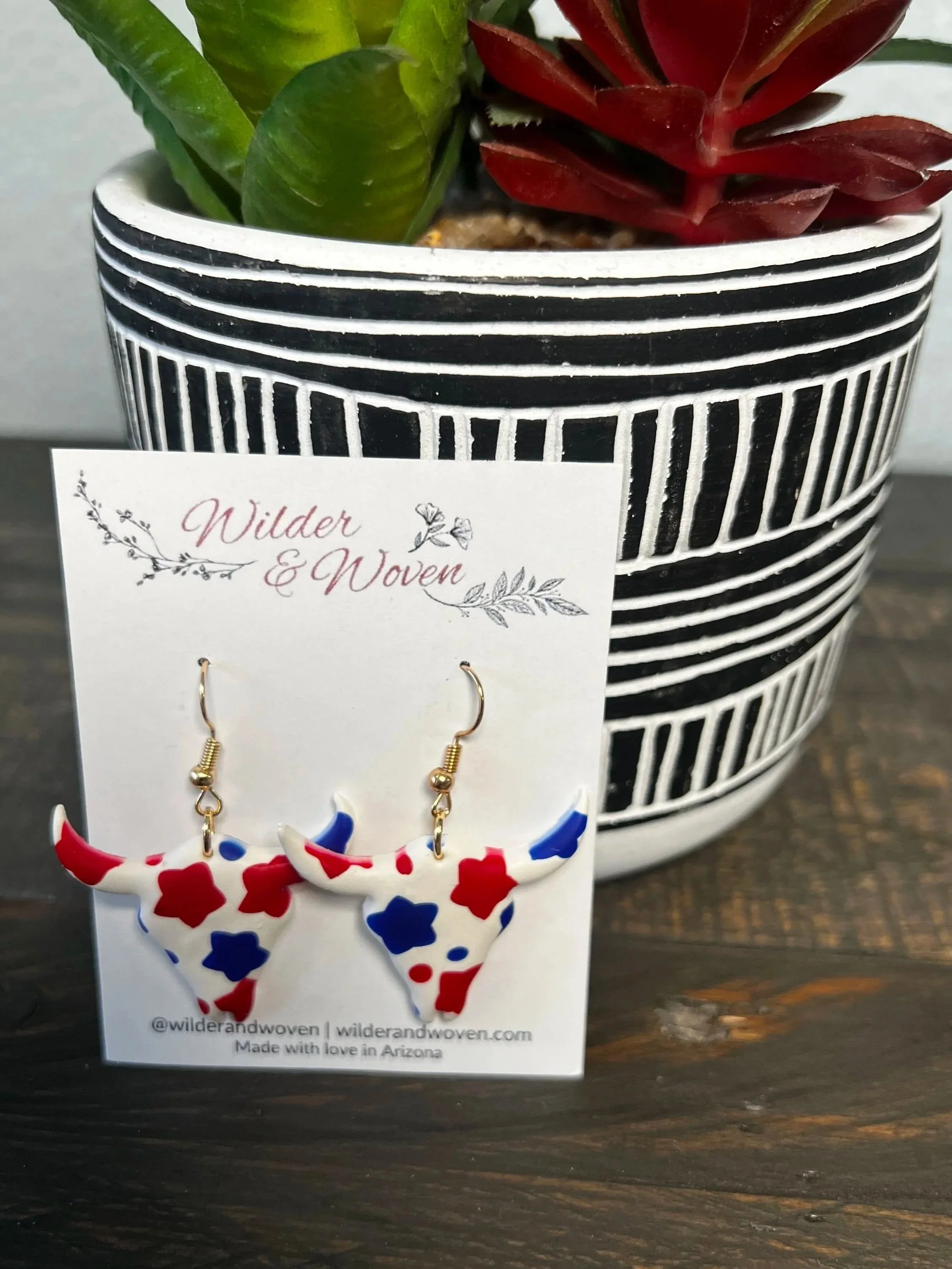 Handcrafted red, white, and blue polymer clay longhorn earrings with patriotic star pattern hanging from gold hooks on a Wilder & Woven display card.