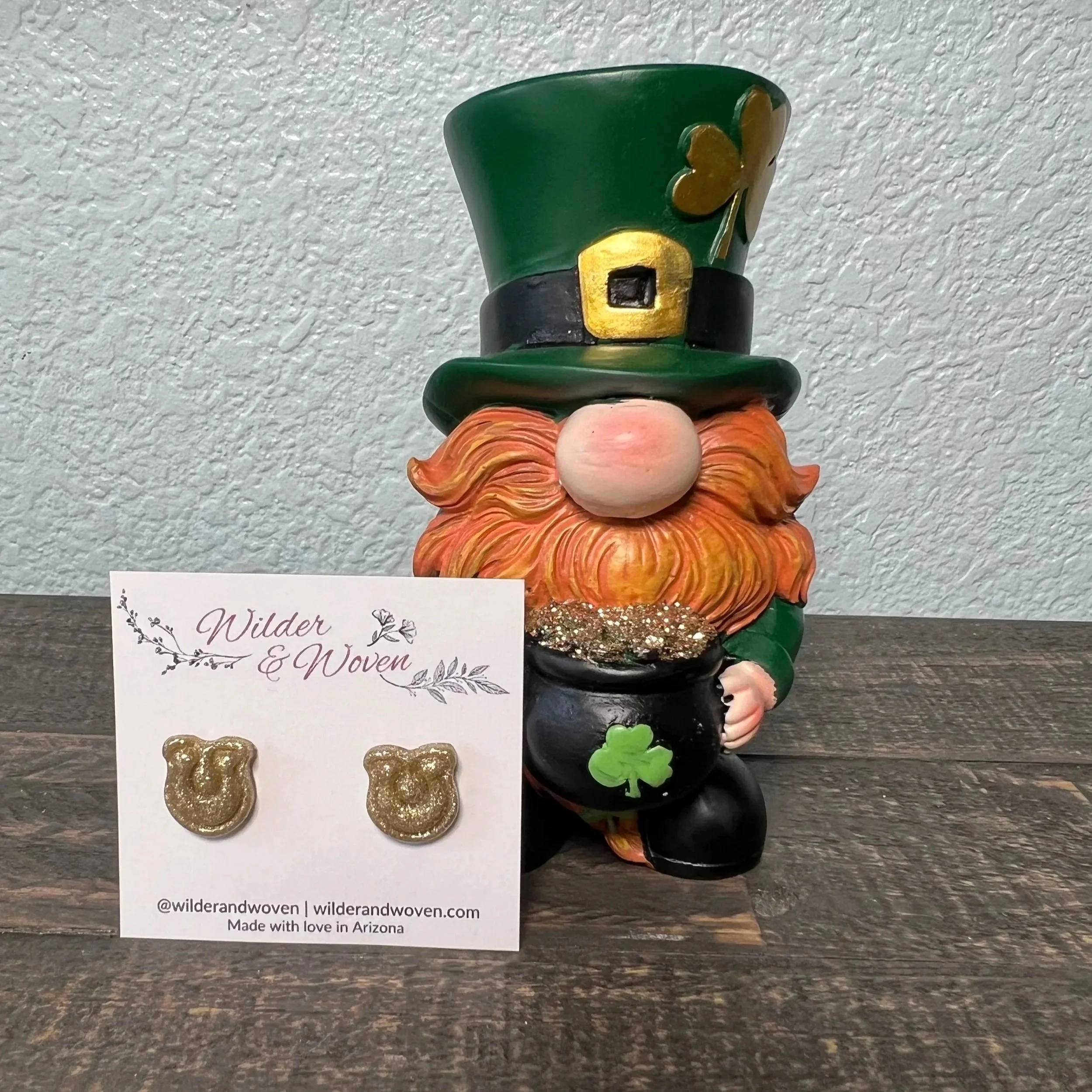 Handmade glittery gold horseshoe stud earrings displayed on a Wilder & Woven jewelry card beside a St. Patrick’s Day leprechaun decoration.
