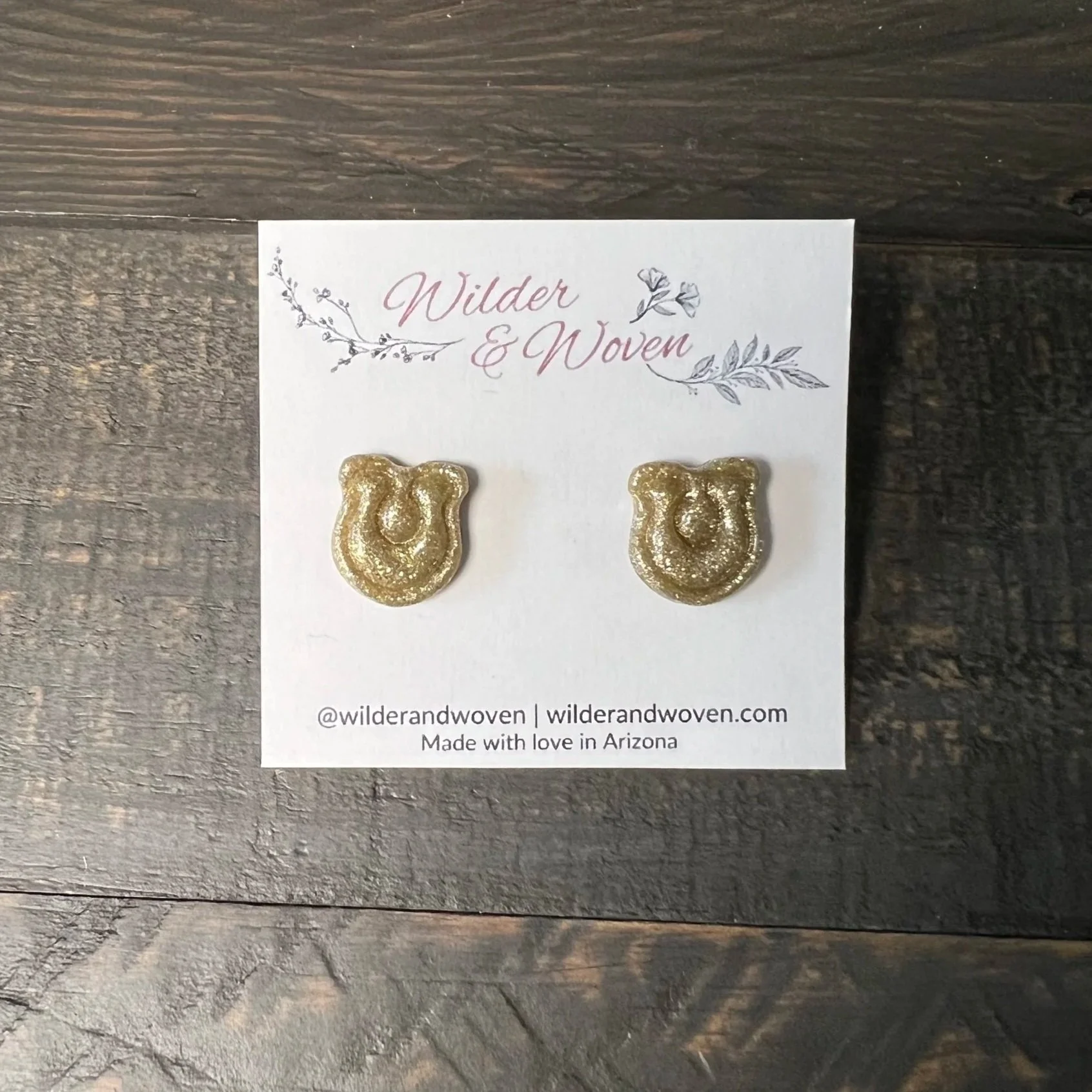 Handmade glittery gold horseshoe stud earrings displayed on a Wilder & Woven jewelry card on a wooden table.