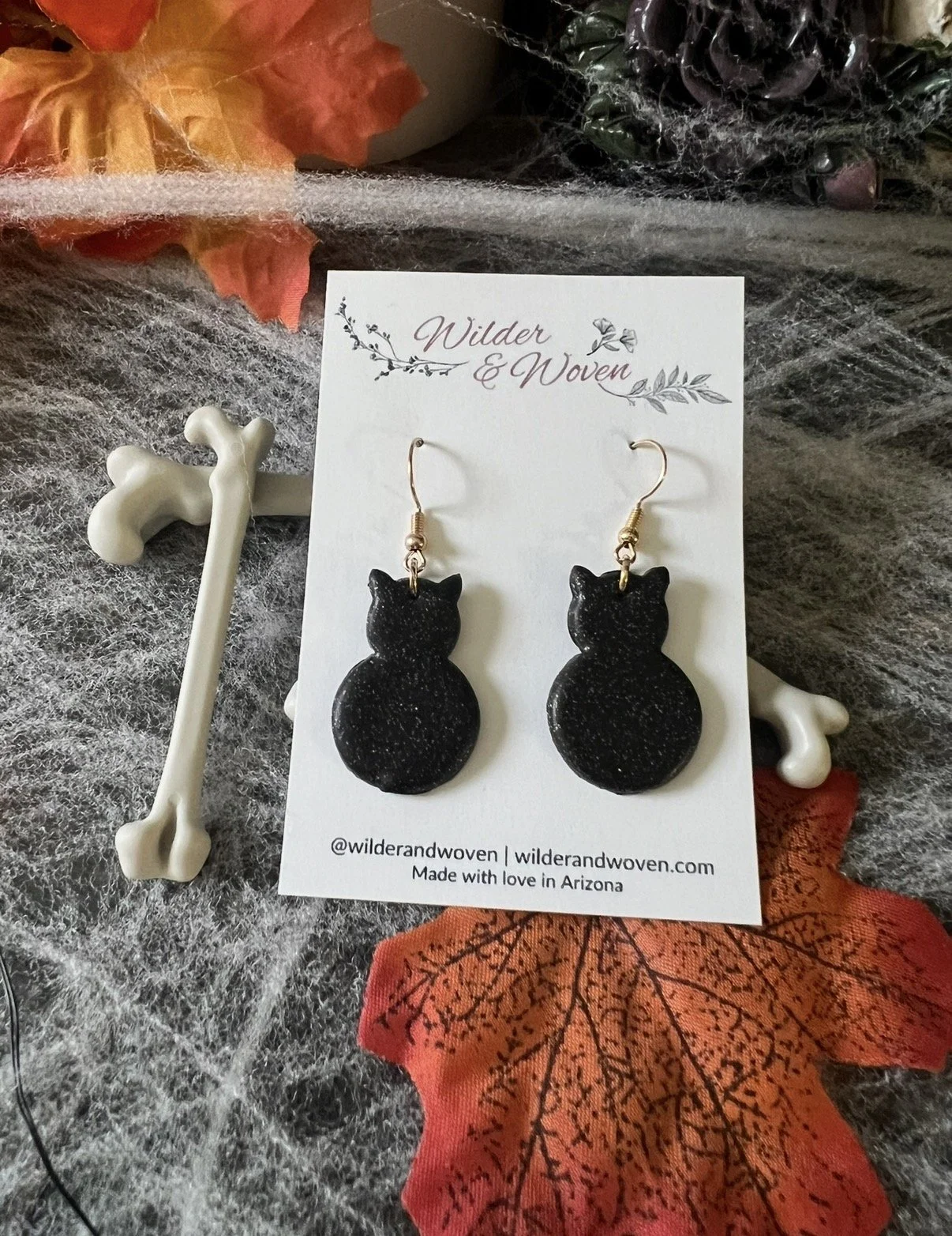 Sparkly black cat polymer clay earrings with gold fishhooks displayed on a Wilder & Woven earring card with Halloween decor.