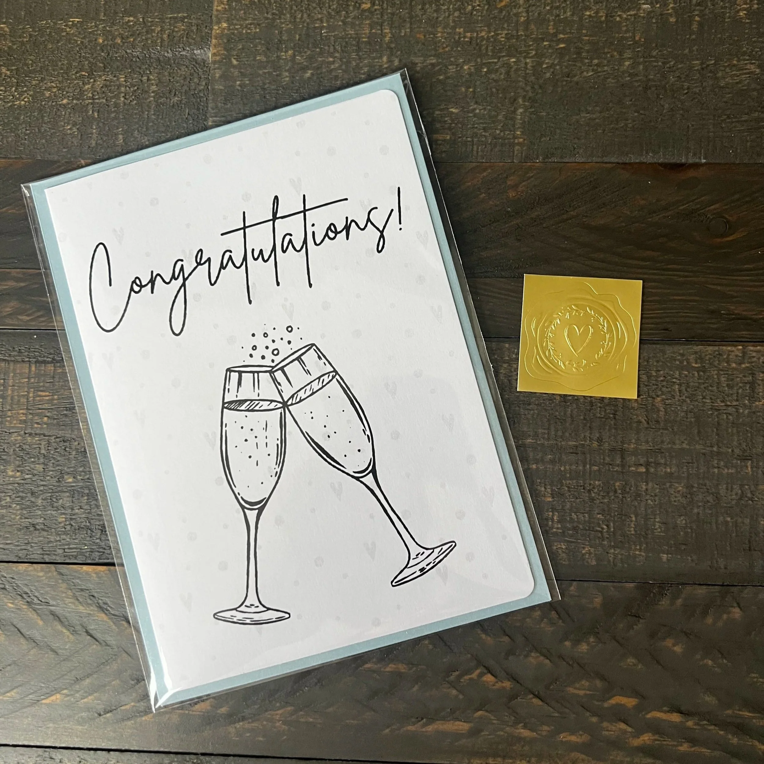 Styled flat lay of Cheers to Happily Ever After Wedding Card with coordinating soft blue envelope and gold wax seal style sticker.