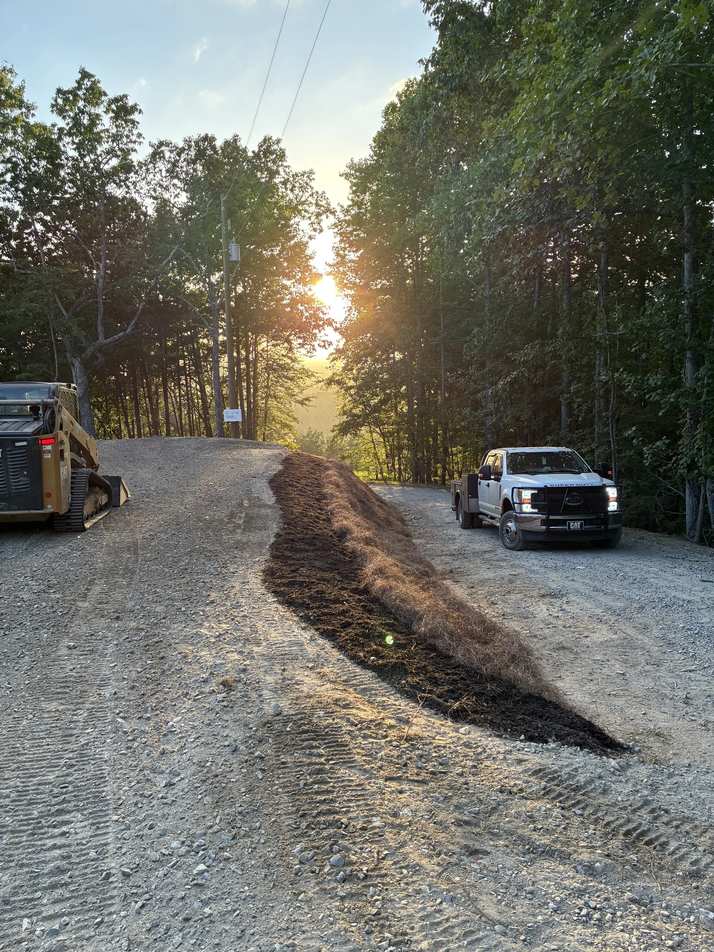A dirt road surrounded by trees, with construction vehicles parked on the sides. The sun is setting in the background through the trees.