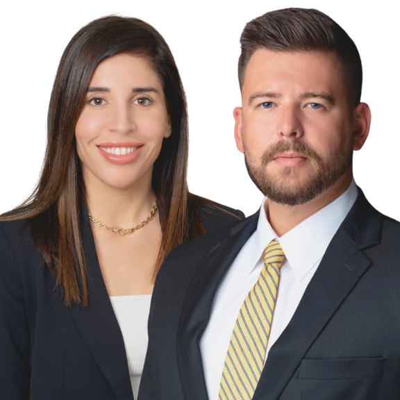 A woman with brown hair wearing a black blazer and white top, smiling, standing next to a man with dark hair and beard, wearing a dark blazer, white shirt, and striped yellow tie, both against a white background.