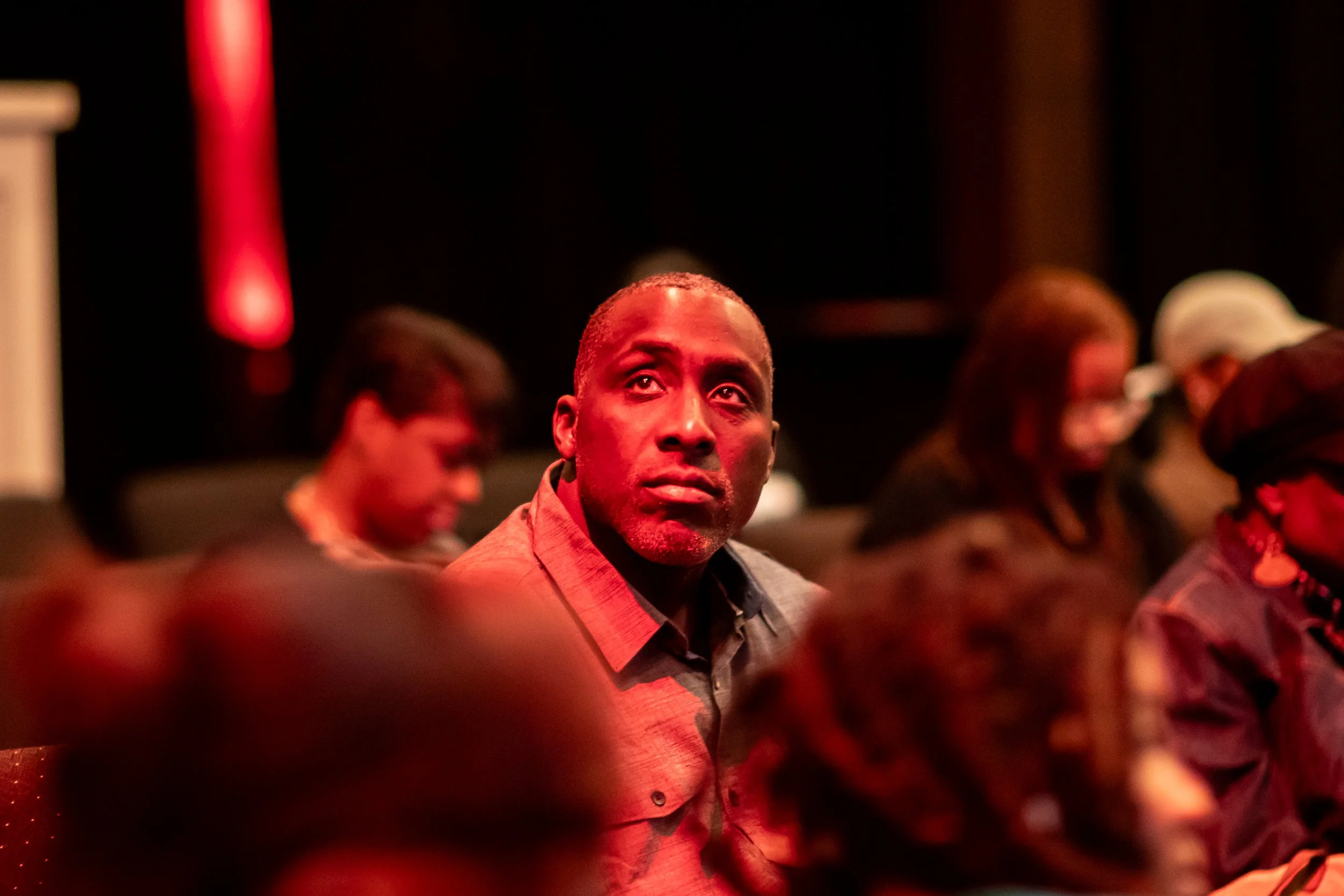 A man sitting in a dark room with red lighting, looking towards the camera, surrounded by other seated people during an event or gathering.
