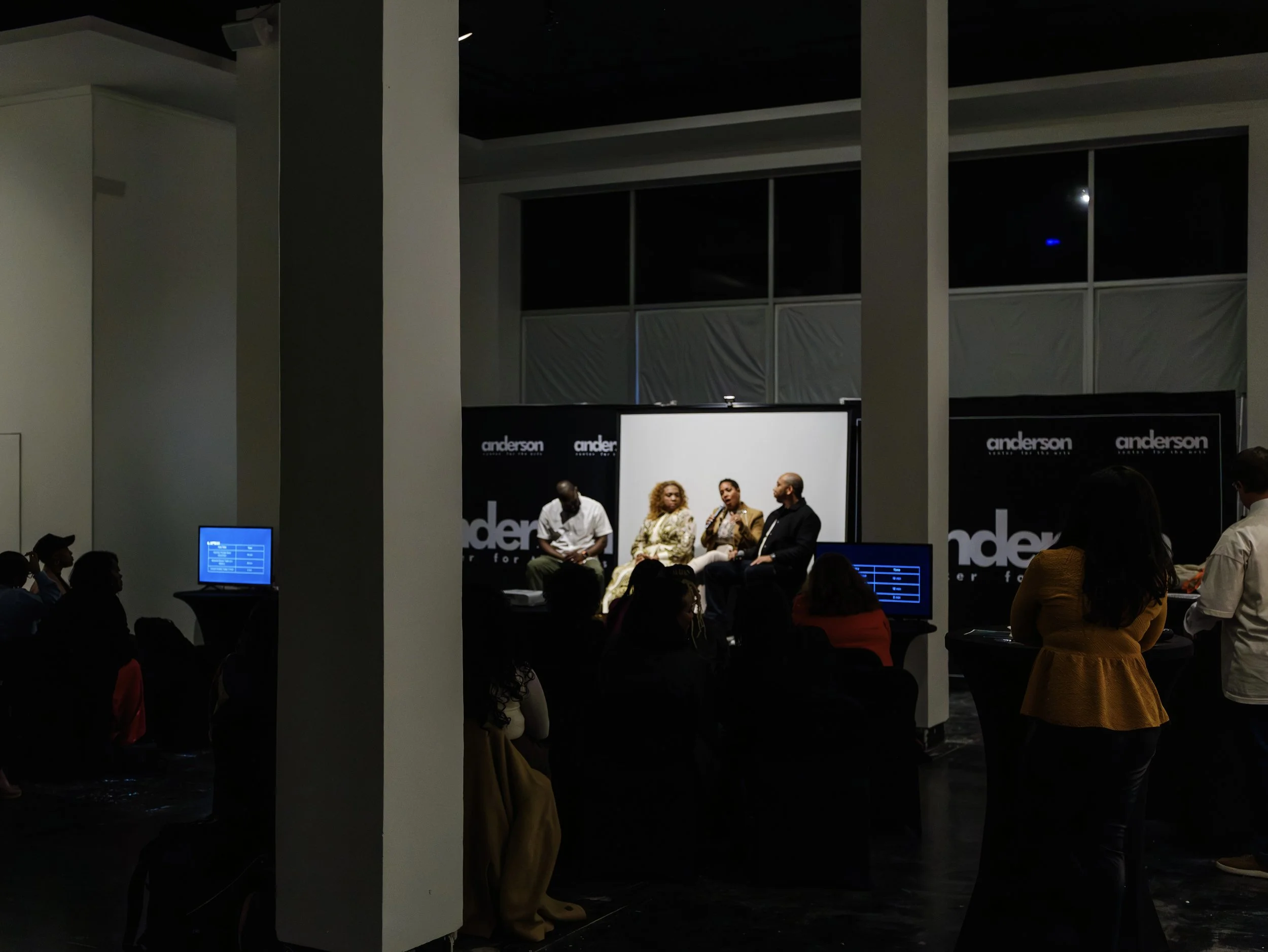 Panel discussion at an event with four people on stage, audience seated in front, screens displaying blue text, and branded black backdrops with white Anderson logo.