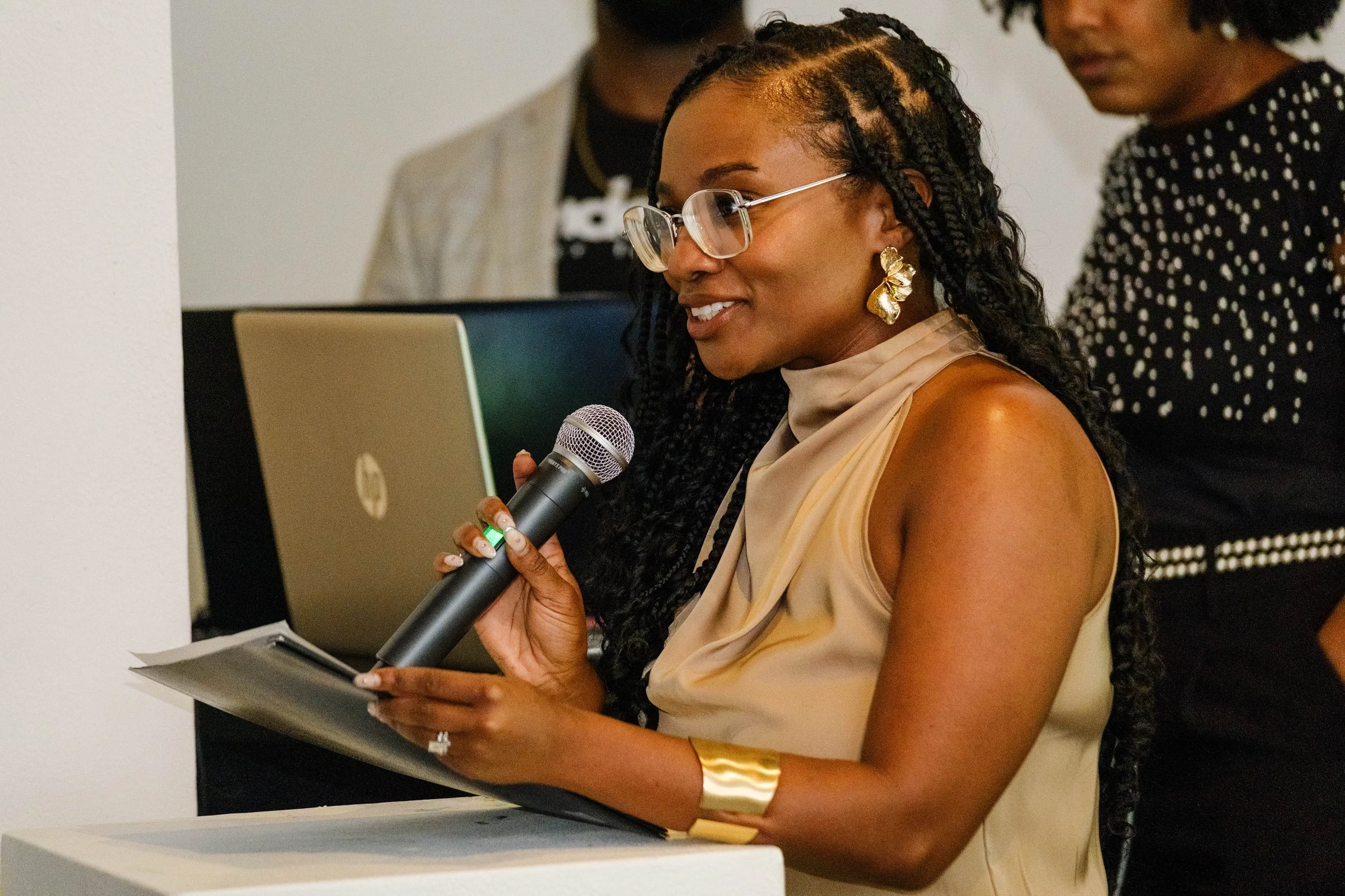 Woman with glasses and gold jewelry speaking into a microphone, holding papers, in a professional setting with others in the background.