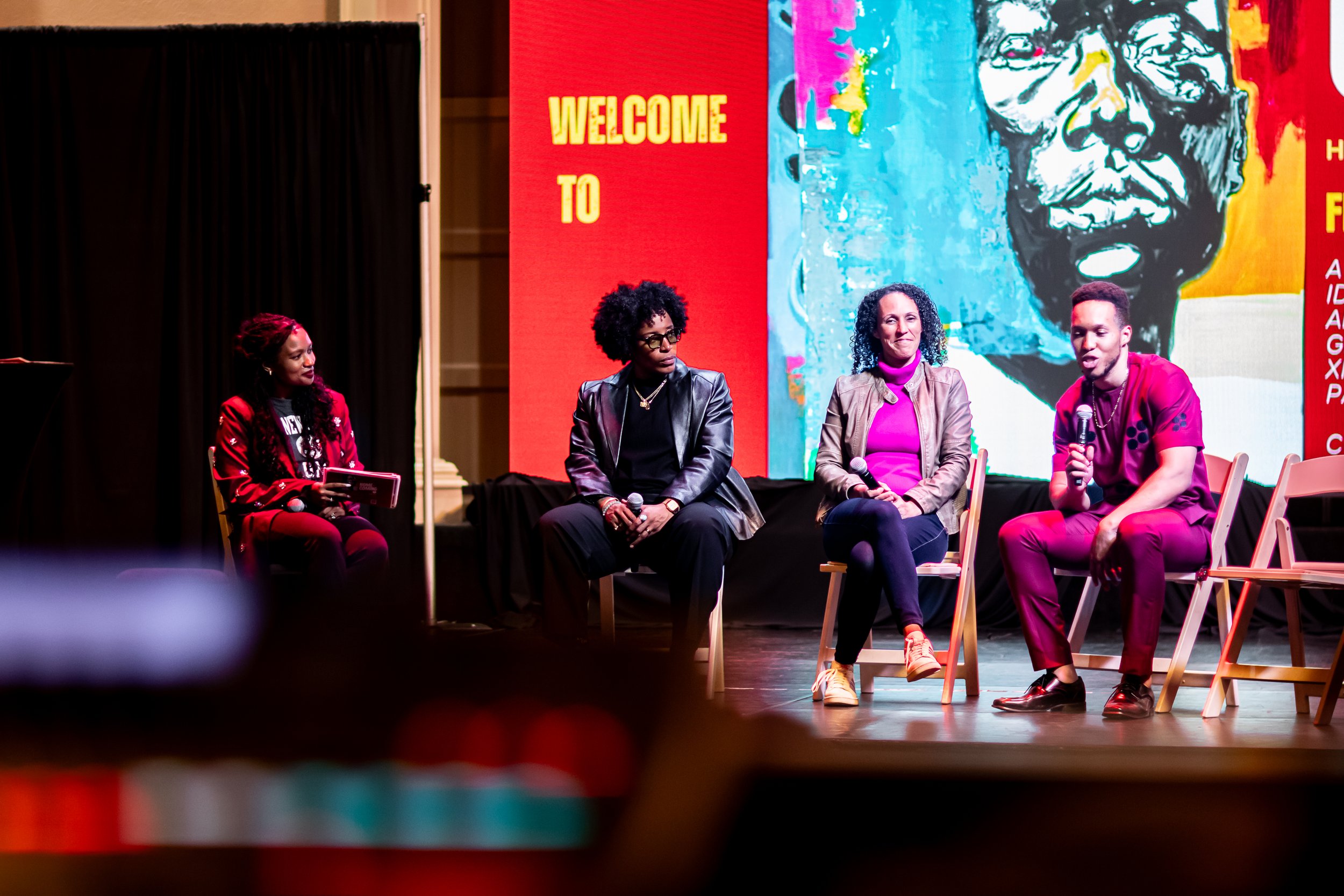 A panel discussion with four people seated on stage, holding microphones, in front of a large colorful mural and red backdrop with yellow text 'Welcome to'.