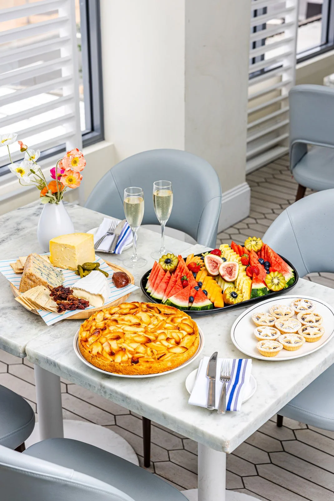 A marble table with a variety of desserts, fruits, cheese, and two glasses of champagne, set in a bright room with large windows and gray chairs.