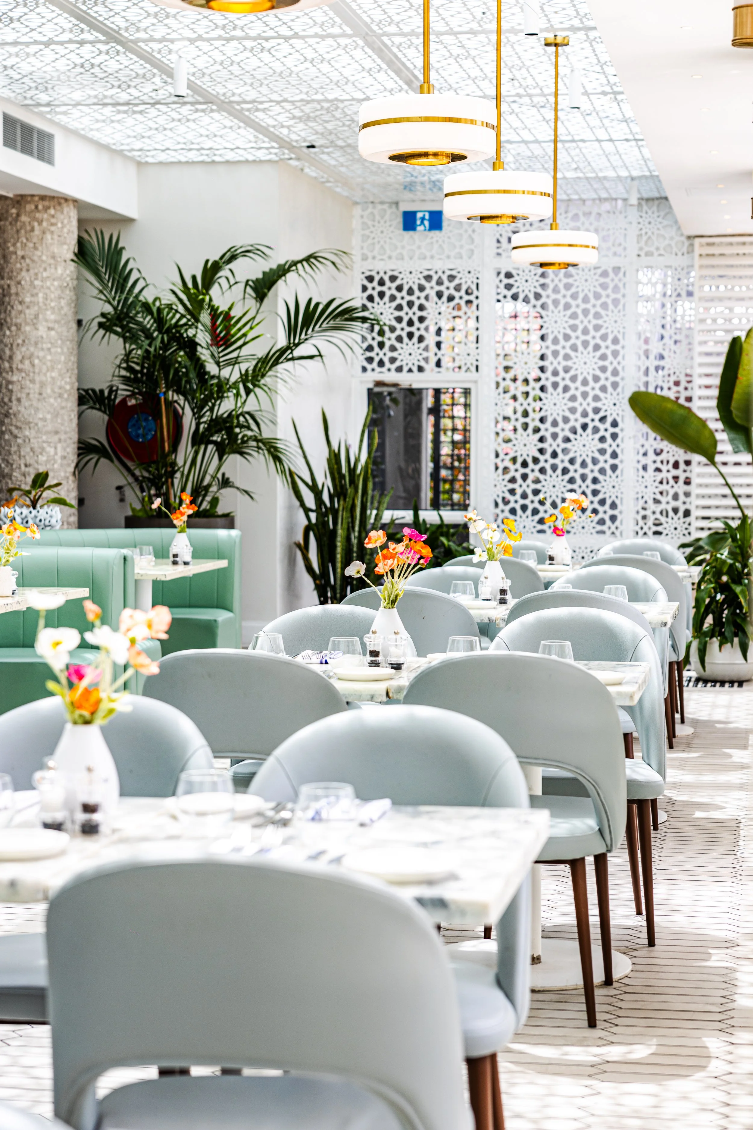 Elegant restaurant interior with white marble tables, light blue chairs, colorful flower arrangements, lush green plants, decorative lighting fixtures, and intricate white lattice walls.