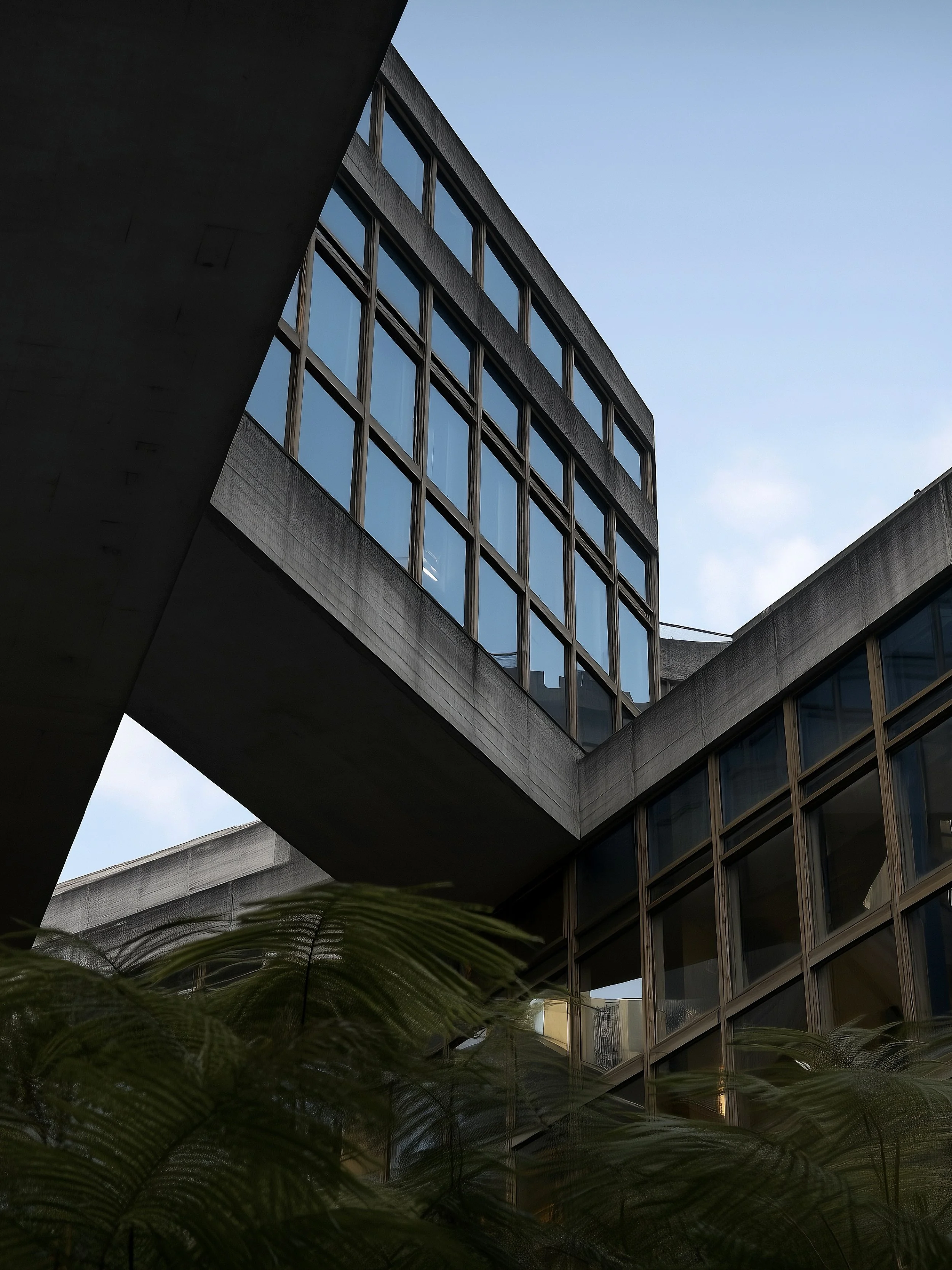 Modern building with glass windows and concrete structure against a clear sky, with foliage at the bottom.