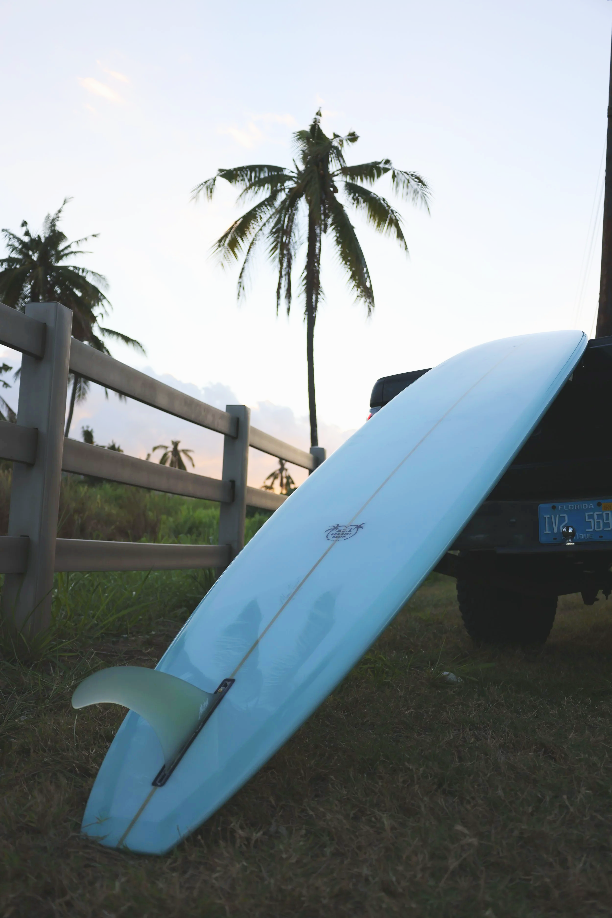 A surfboard leaning against a vehicle near palm trees at sunset or sunrise in a tropical setting.