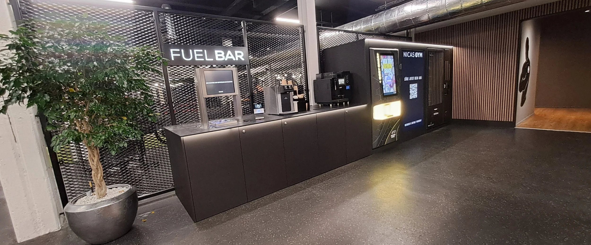 Ein Getränke- und Kaffeeautomat, den Fuel Bar, an der Wand mit einem großen grünen Pflanzkübel daneben. Es gibt auch eine andere Maschine für Kaffee und ein digitales Menü. Im Hintergrund sind dunkle Wände und Holzbretter.