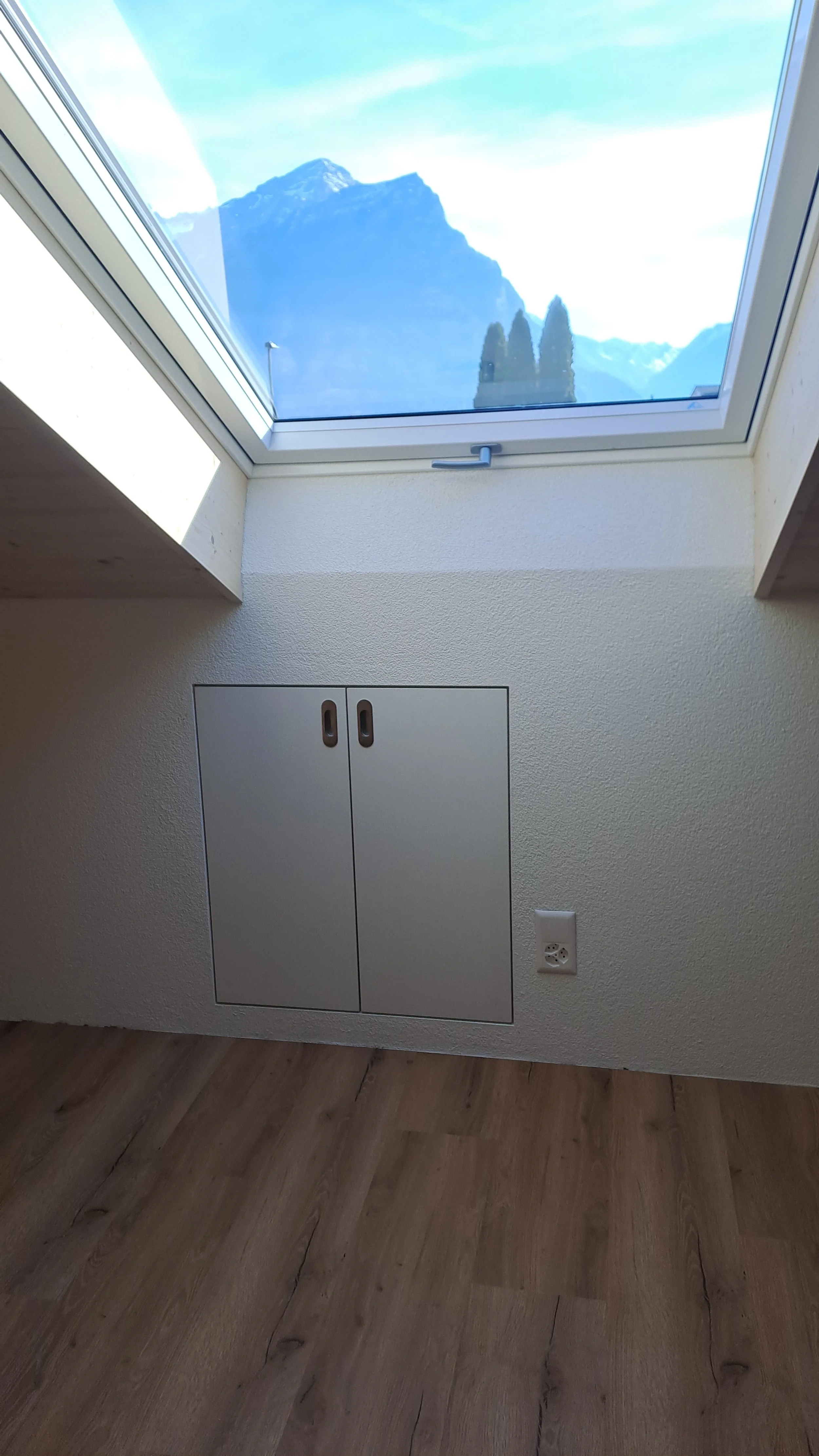 Innenraum mit Dachfenster, Blick auf Berge und Himmel, kleine Tür im Boden, europäischer Stecker an Wand, Holzlaminatboden.