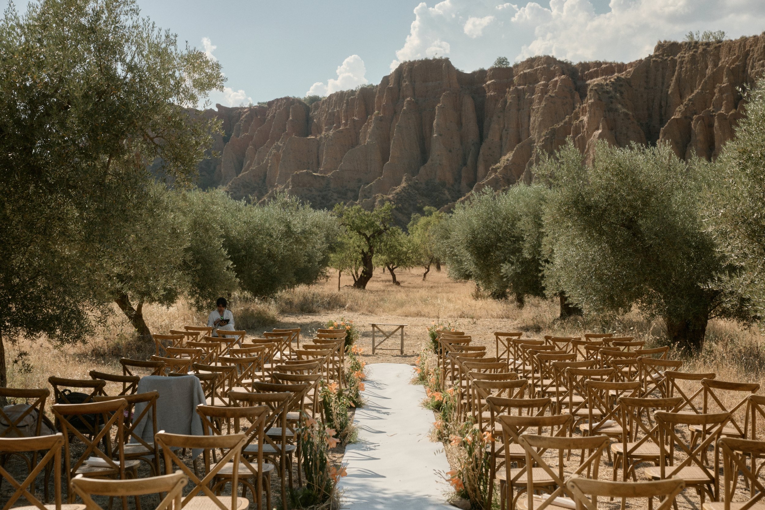 Ein Outdoor-Ambient mit Stühlen und Blumen, vermutlich für eine Trauung, inmitten einer kargen Landschaft mit Bäumen und hohen Felswänden im Hintergrund.