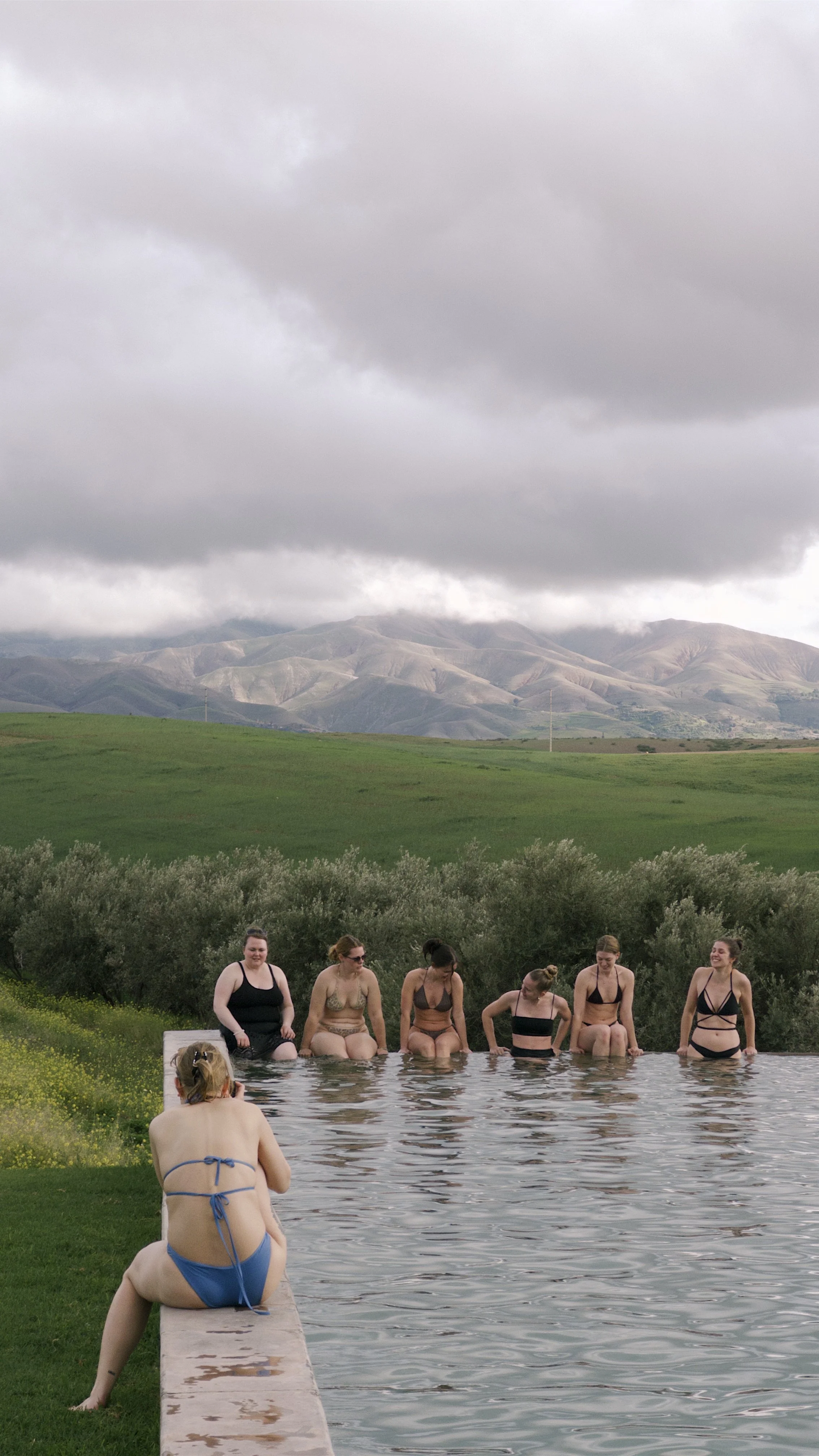 A group of women in swimsuits sitting on the edge of an outdoor pool, with one woman in a blue swimsuit sitting on the edge facing away, holding a camera. The background features green hills, bushes, and cloudy sky. Bachelorette trip morocco.