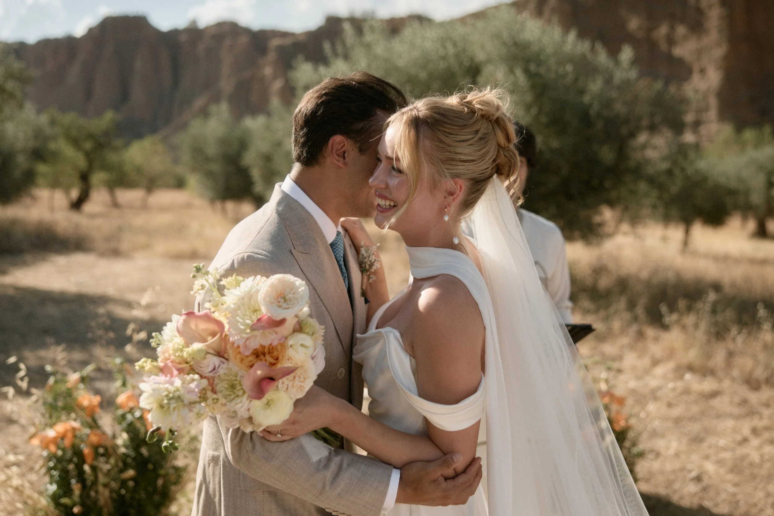 Ein Brautpaar bei einer Hochzeit, umgeben von einer natürlichen Landschaft mit Bäumen und Bergen im Hintergrund.