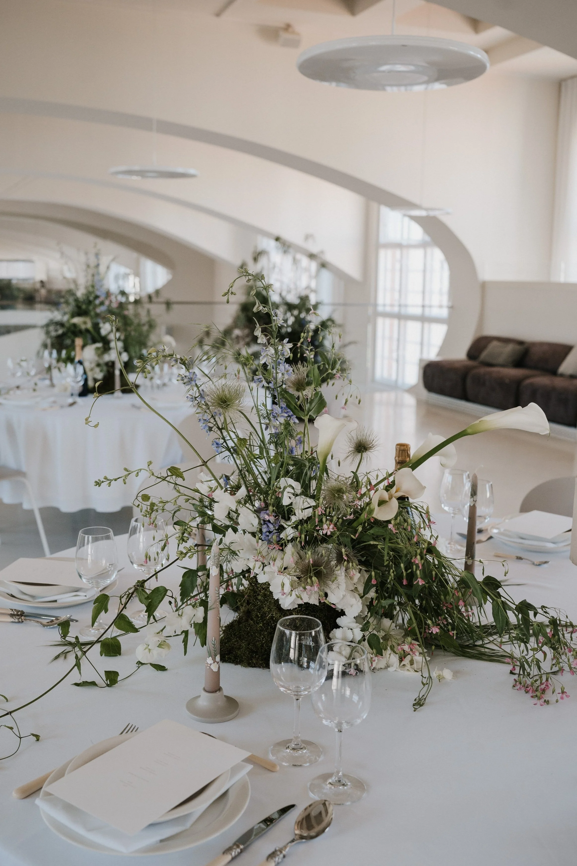 Ein festlich gedeckter Tisch mit einer Blumenmitte aus weißen und grünen Blumen sowie Kerzen in einem hellen Raum.