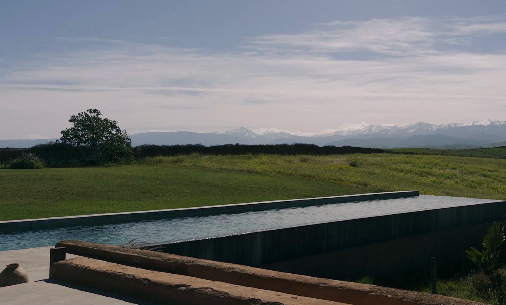 A landscape with a long, narrow swimming pool in the foreground, green fields, trees, mountains, and a cloudy sky in the background.