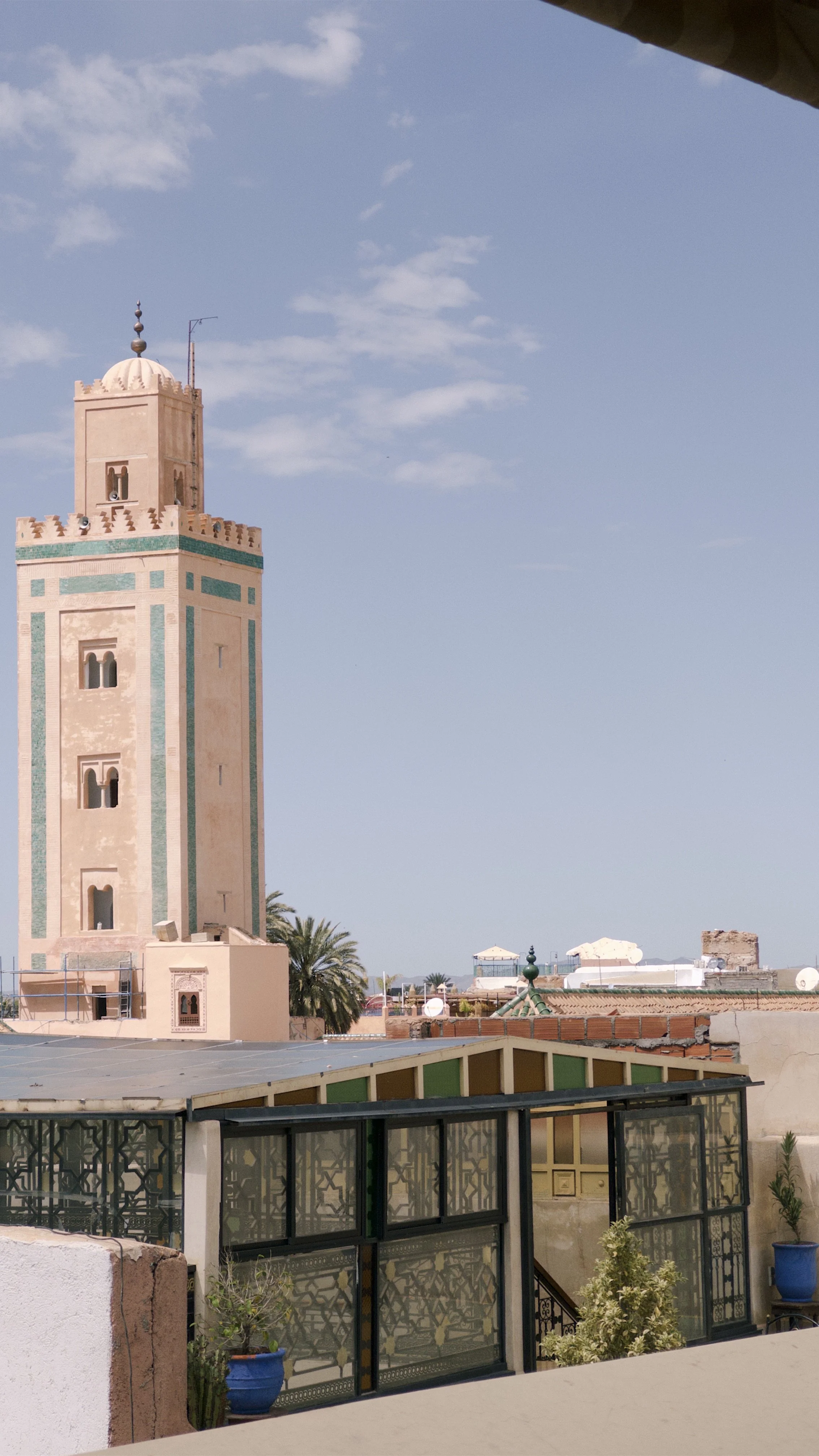 A tall, pastel-colored Moroccan minaret with a blue-green decorative band near the top, set against a bright blue sky with some clouds, surrounded by rooftops, palm trees, and satellite dishes.