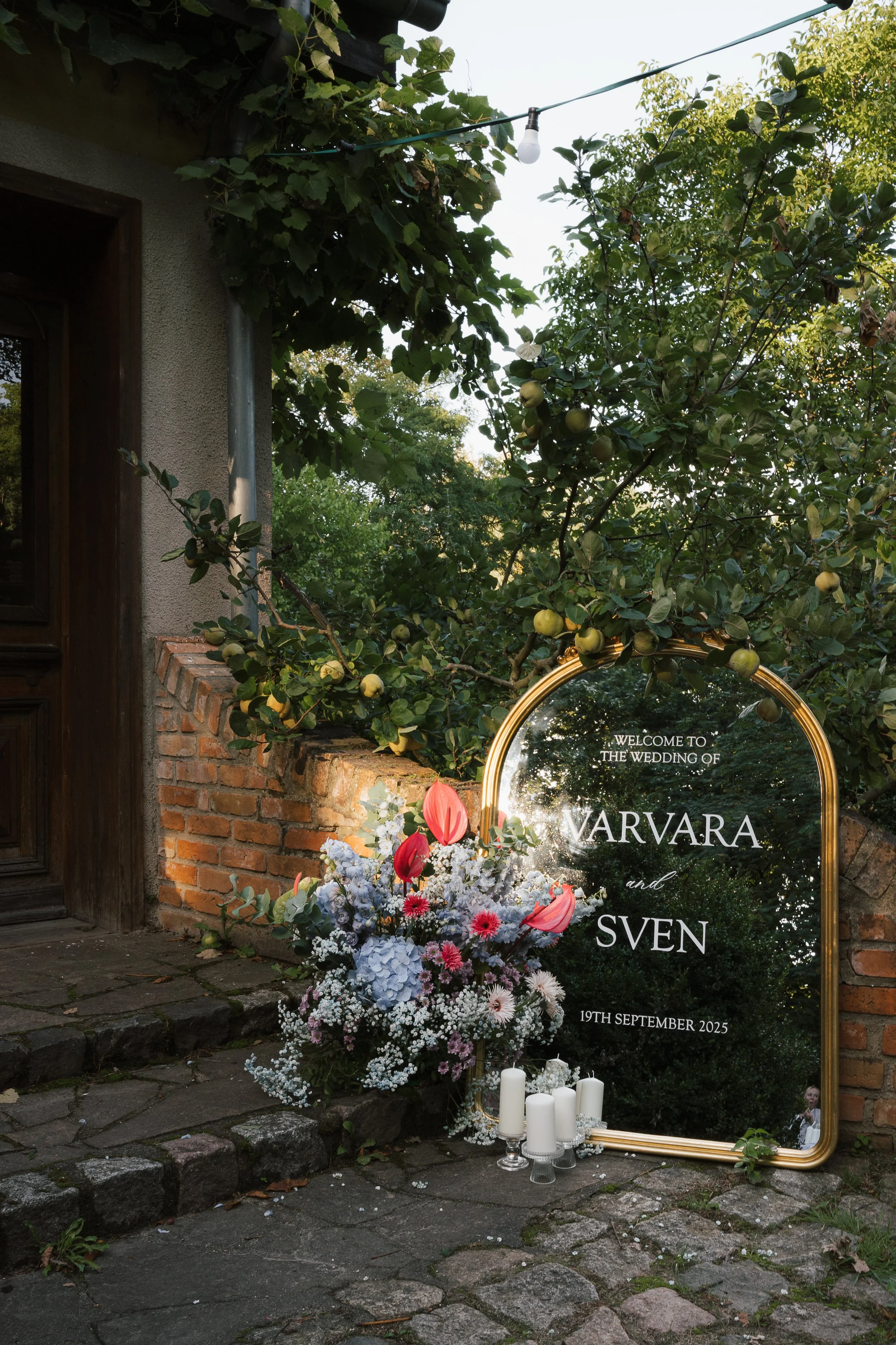 Ein reflektiernder Spiegel mit Goldrahmen, der die Namen des Hochzeitspaares Waldvara und Sven sowie das Datum 19. September 2025 anzeigt, umgeben von einem Blumenarrangement mit weißen, pinken und roten Blumen, Kerzen und grünen Ästen neben einer Steinmauer vor einem Haus, umgeben von Bäumen.