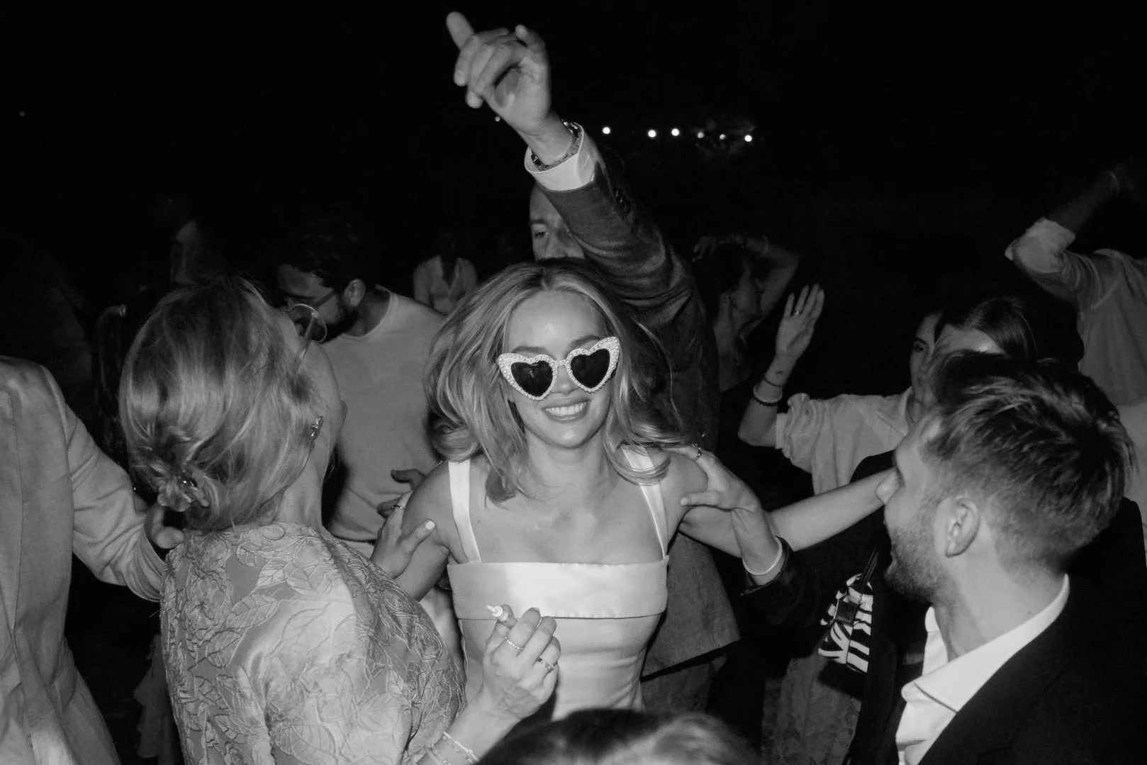 Junge Frau mit Herz-Ohrringen lächelnd auf einer Party, umgeben von tanzenden Menschen in dunkler Umgebung.