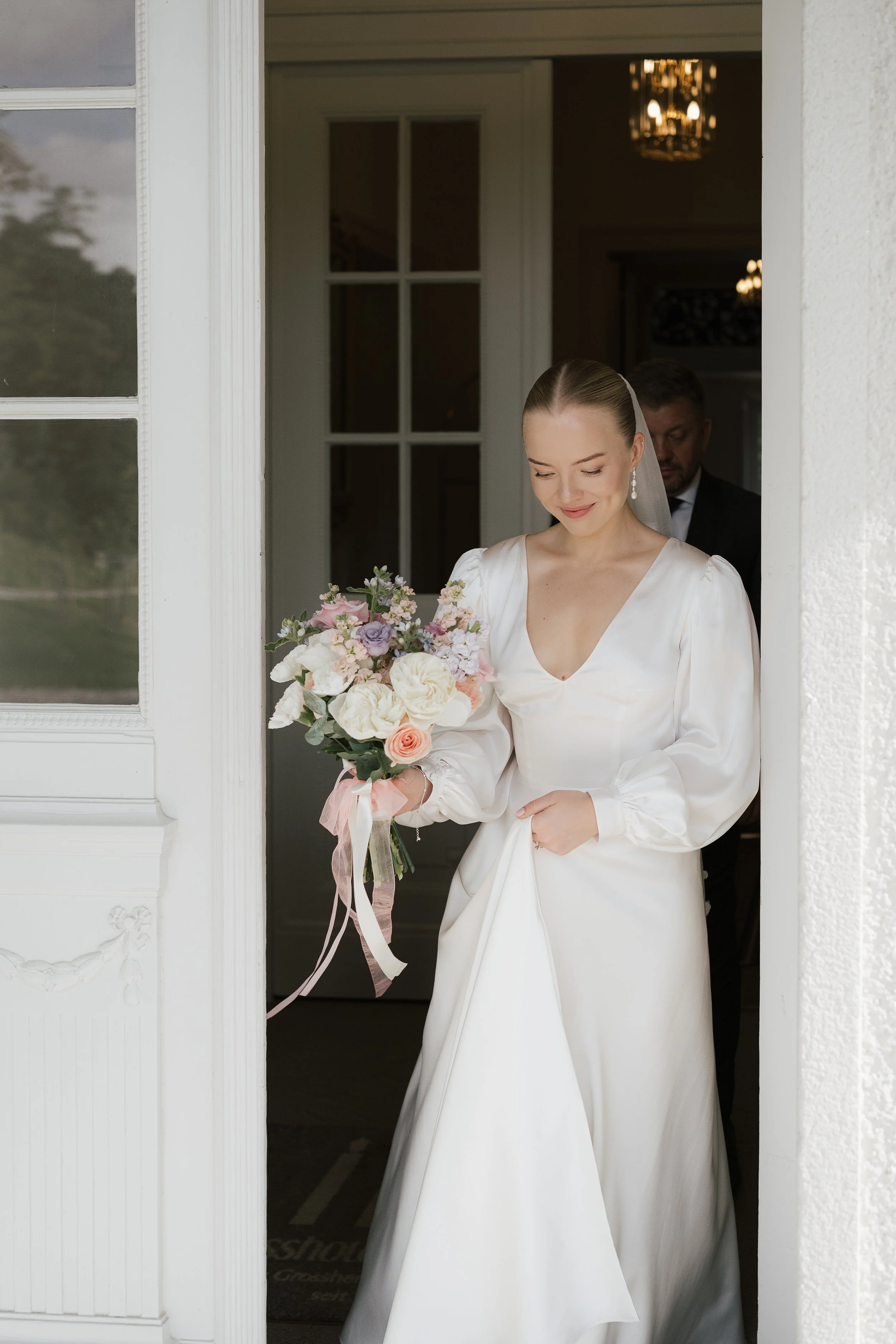 Braut hält einen Blumenstrauß und schaut lächelnd nach unten, während sie ihre Brautkleid anhebt, im Türrahmen stehend, im Hintergrund ein Mann in dunklem Anzug.