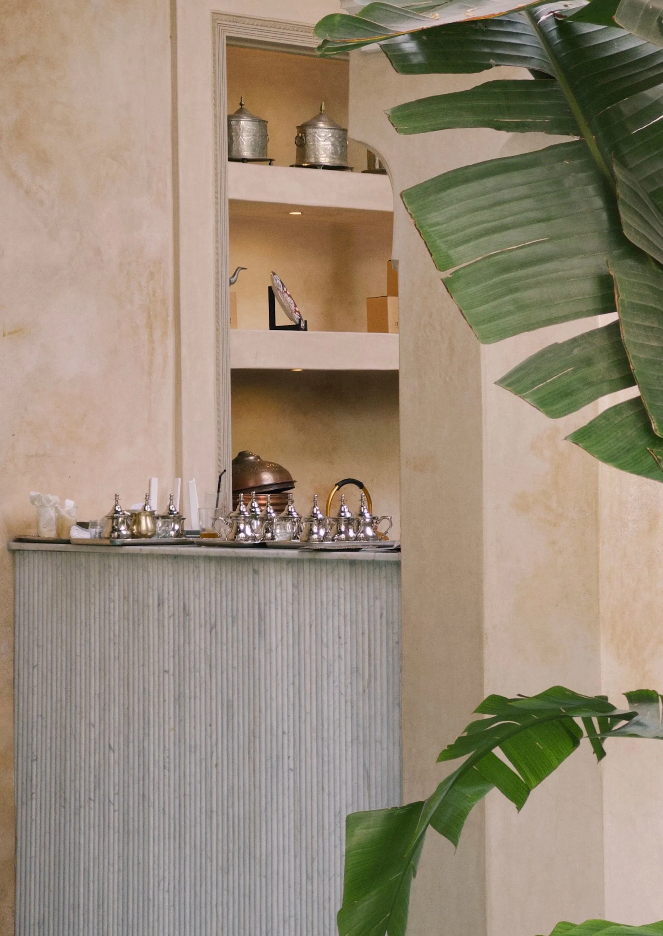 A neutral-toned reception desk with metal teapots and decorative items, partially obscured by large green tropical leaves in the foreground.