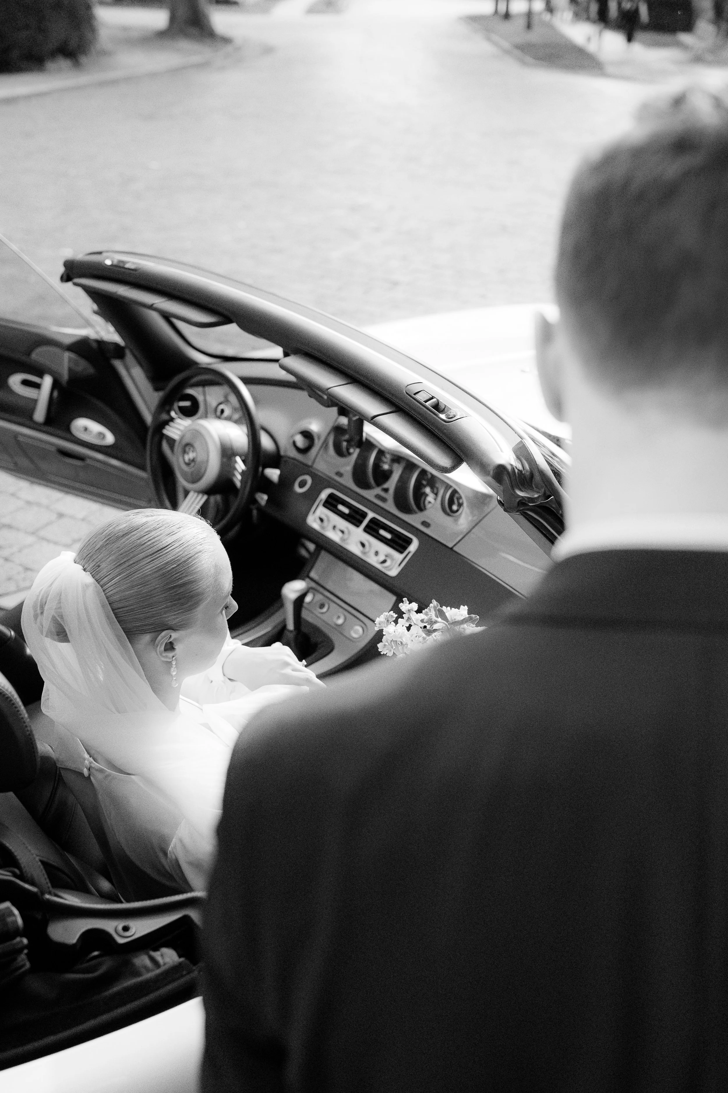 Braut und Bräutigam sitzen in einem Auto, möglicherweise an ihrem Hochzeitstag. Die Braut trägt einen Schleier und ein weißes Kleid, während der Bräutigam einen Anzug trägt. Das Bild ist in Schwarz-Weiß aufgenommen.