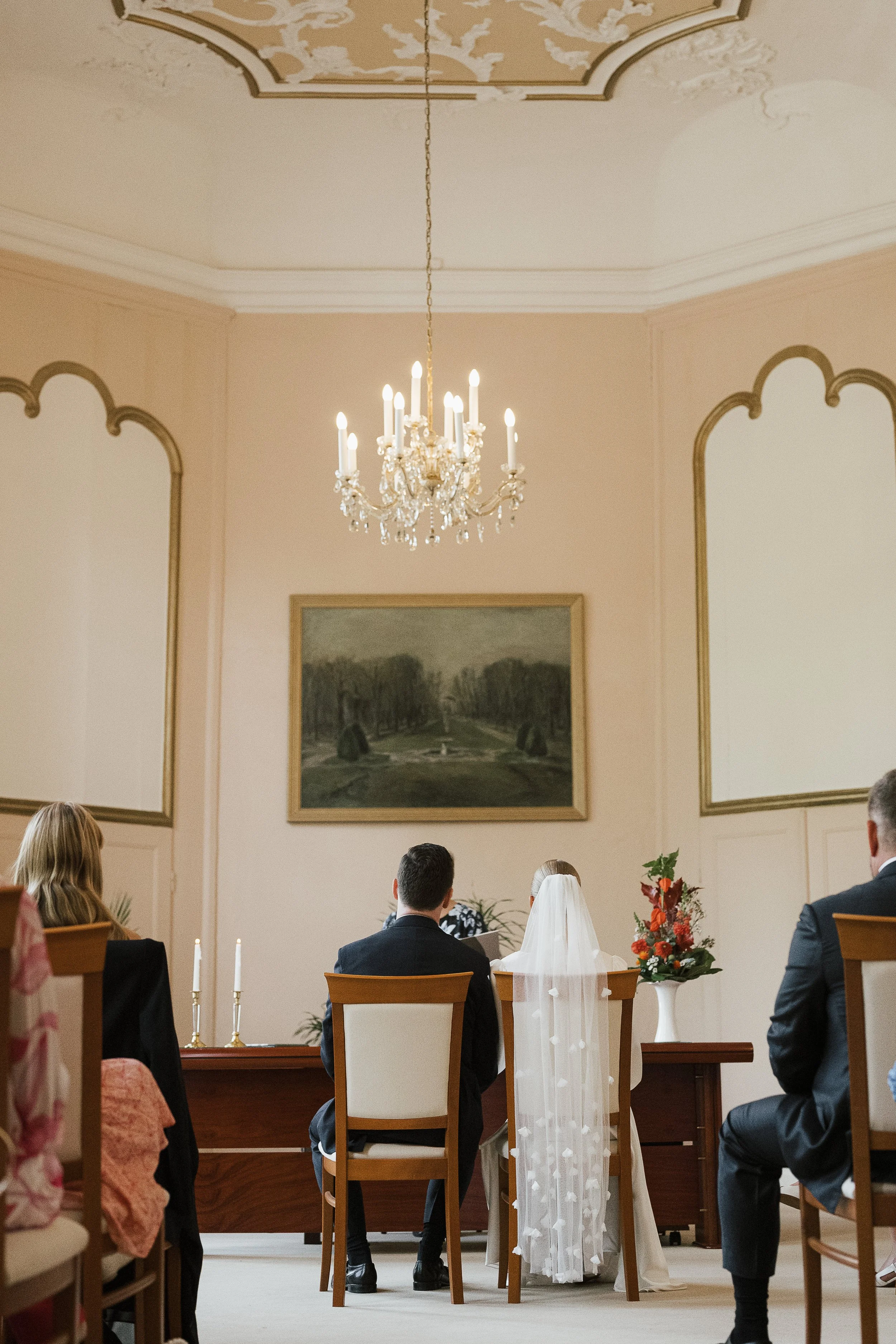 Eine Hochzeit im Innenraum mit einer Braut in weißem Kleid und Schleier, die vor einem Predigtstuhl sitzt, umgeben von Gästen, in einem elegant dekorierten Raum mit Kristallleuchter und Landschaftsbild an der Wand.