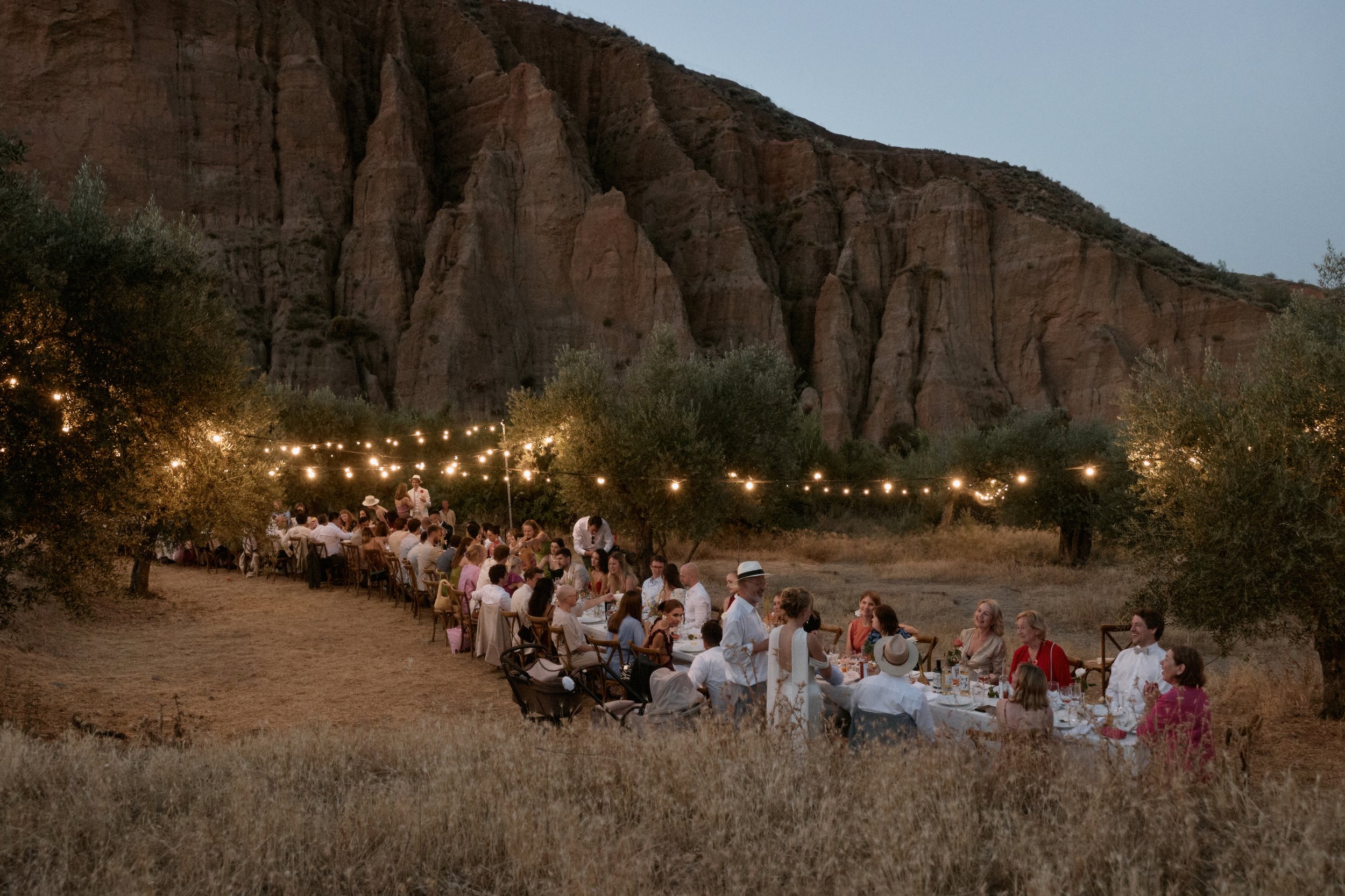 Eine große Gruppe Menschen sitzt an einem langen Esstisch im Freien, unter Lichterketten, mit Bergen im Hintergrund, abends.