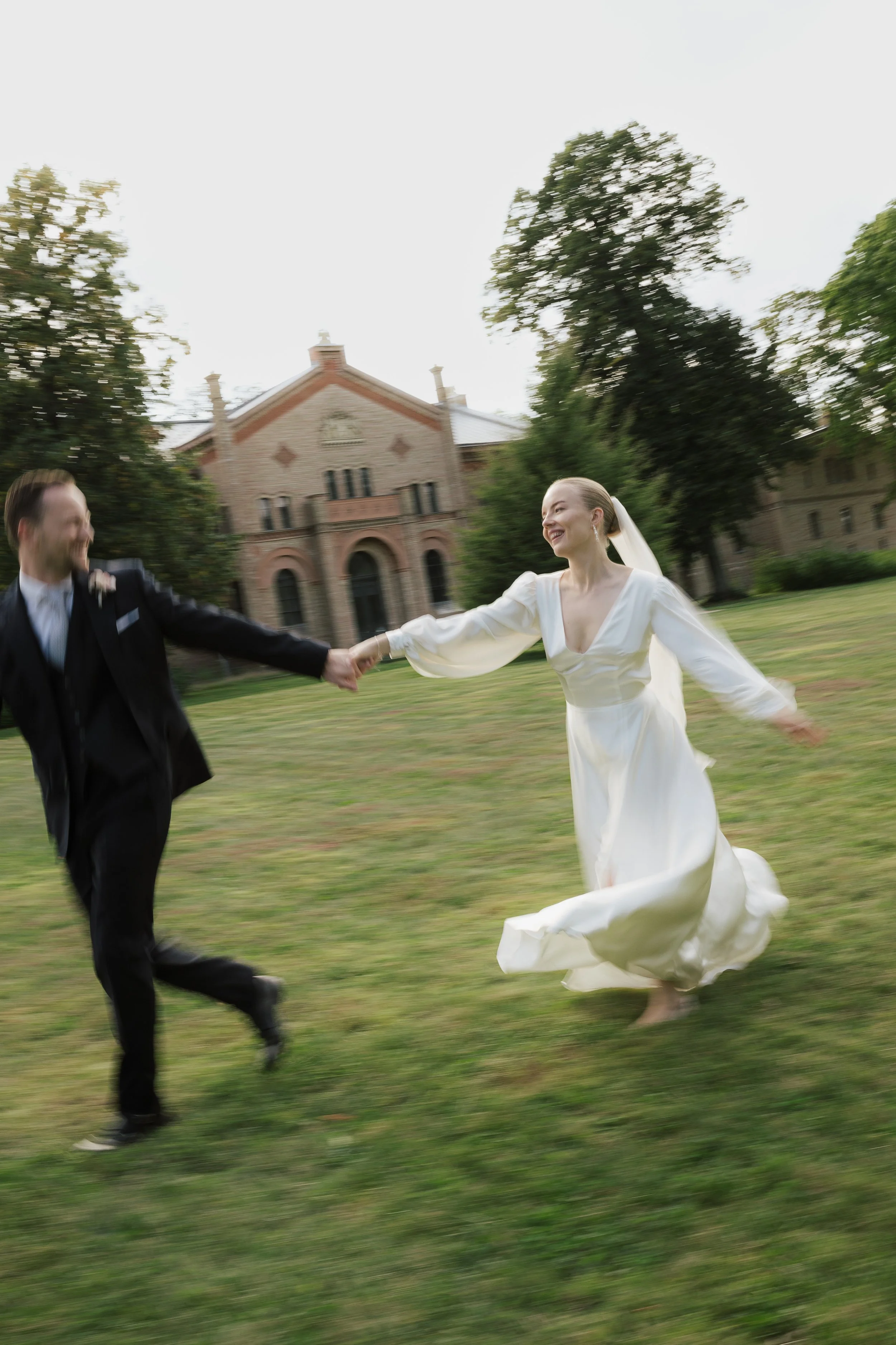 Ein Hochzeitspaar läuft lachend über eine Wiese, die Braut trägt ein weißes Kleid und der Bräutigam einen schwarzen Anzug.