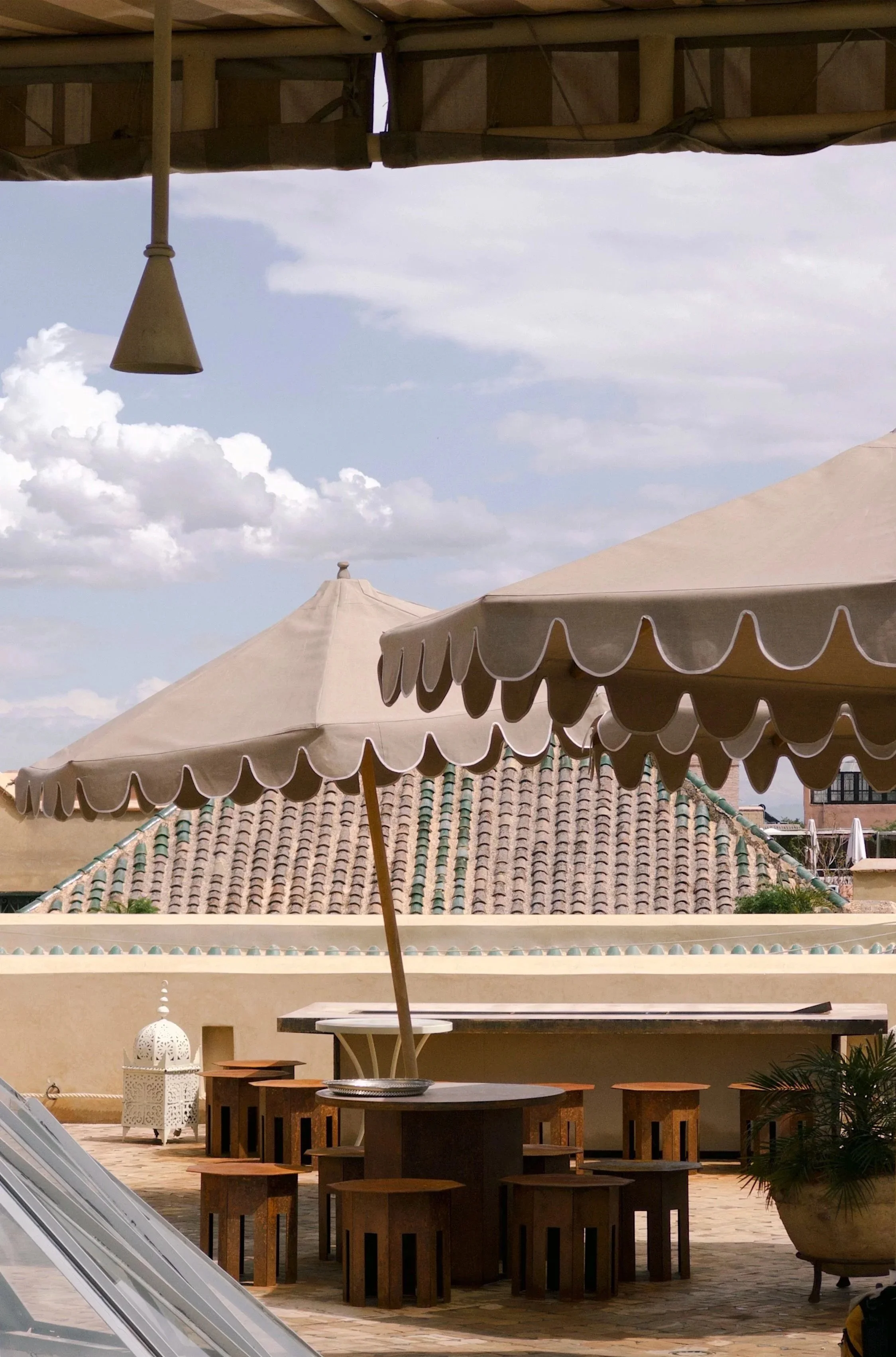 Outdoor patio area with round table under a large beige umbrella, surrounded by wooden stools, with another umbrella in the background and rooftop tiles visible, under a partly cloudy sky.