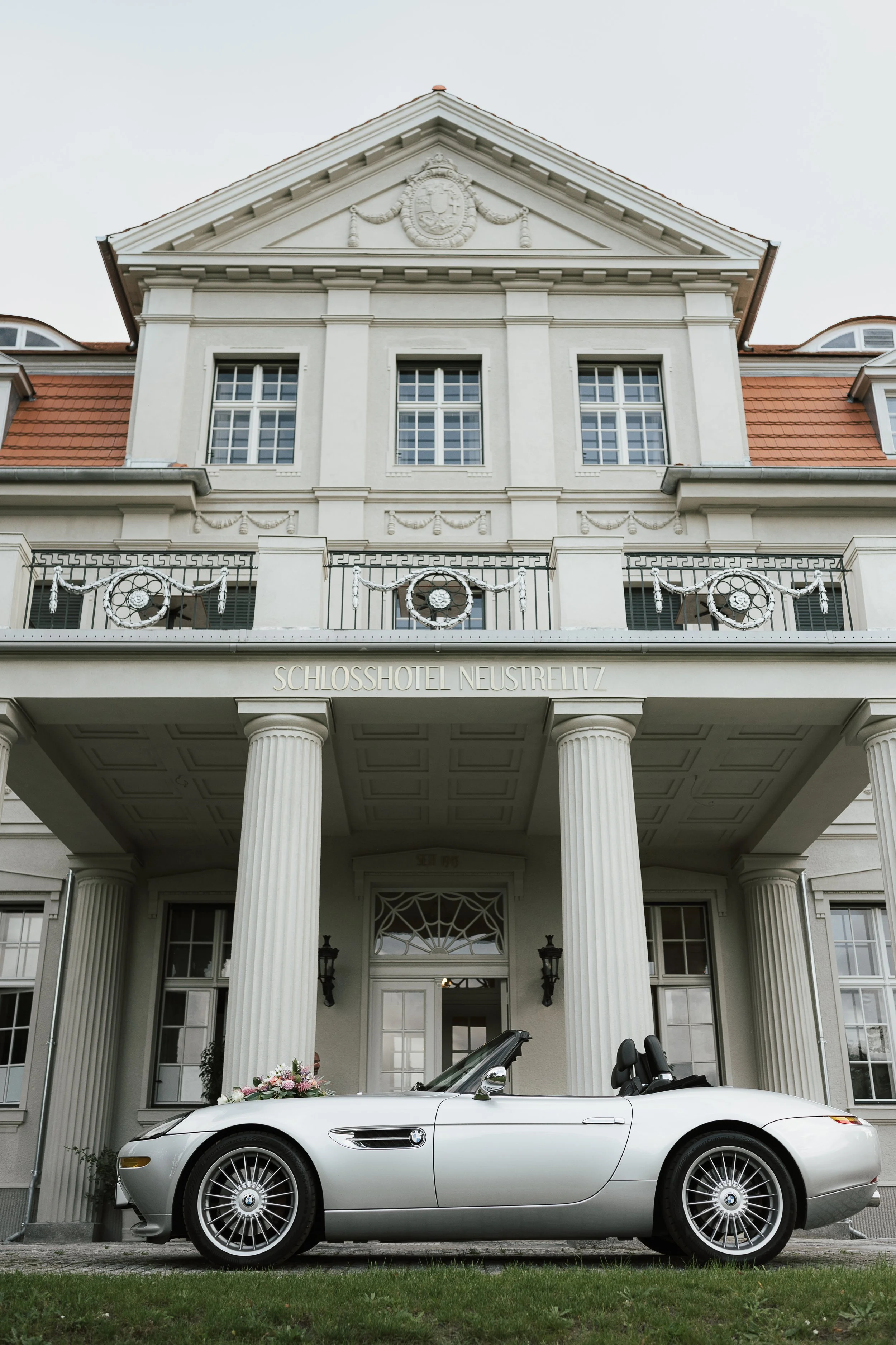 Ein weißer Cabriowagen, verziert mit Blumen, steht vor dem Schlosshotel Neustrelitz, das eine neoklassizistische Fassade mit Säulen hat.