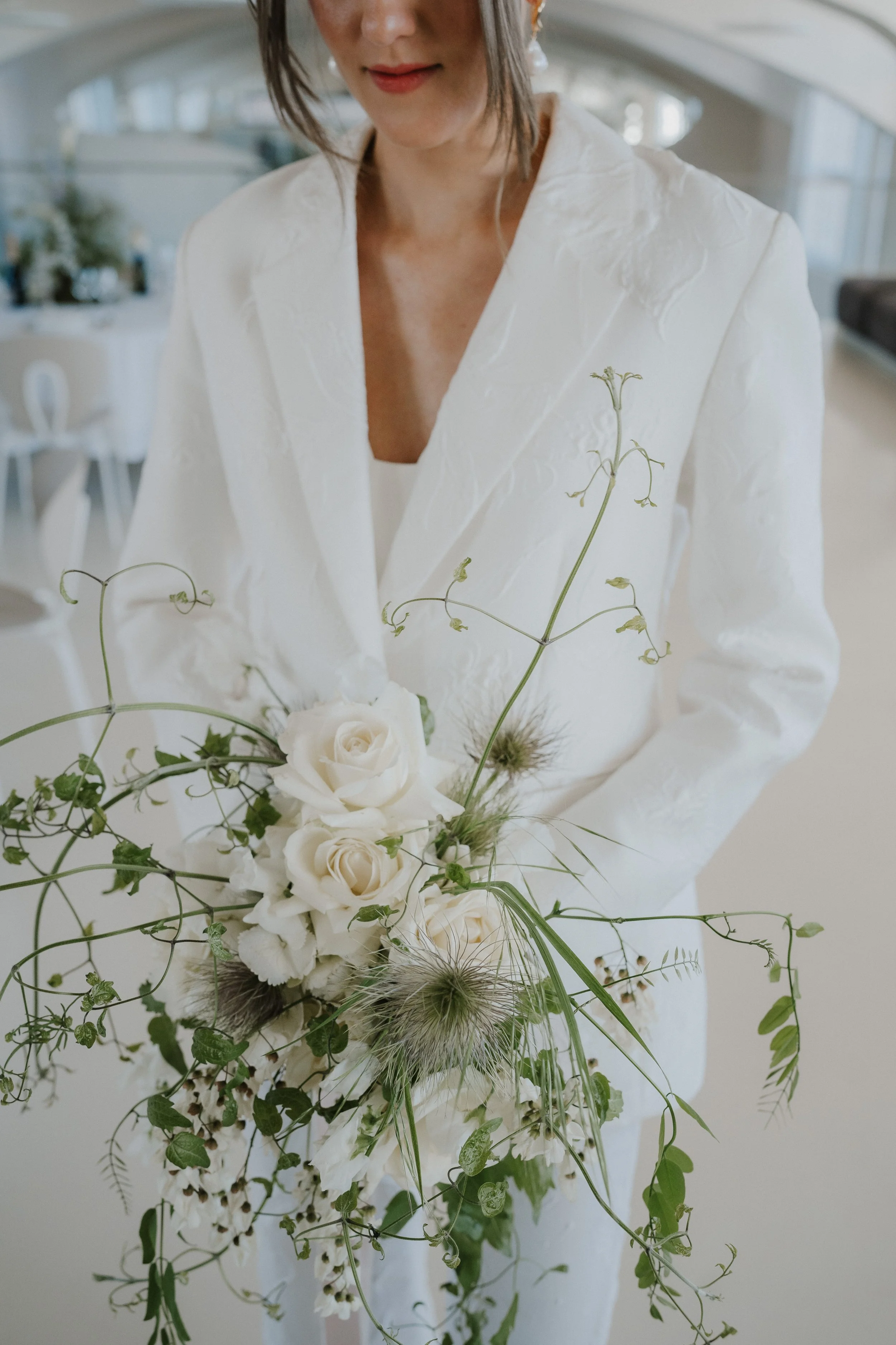 Eine Frau in weißem Anzug hält einen weißen Blumenstrauß mit Rosen, Gräsern und zarten Blüten.