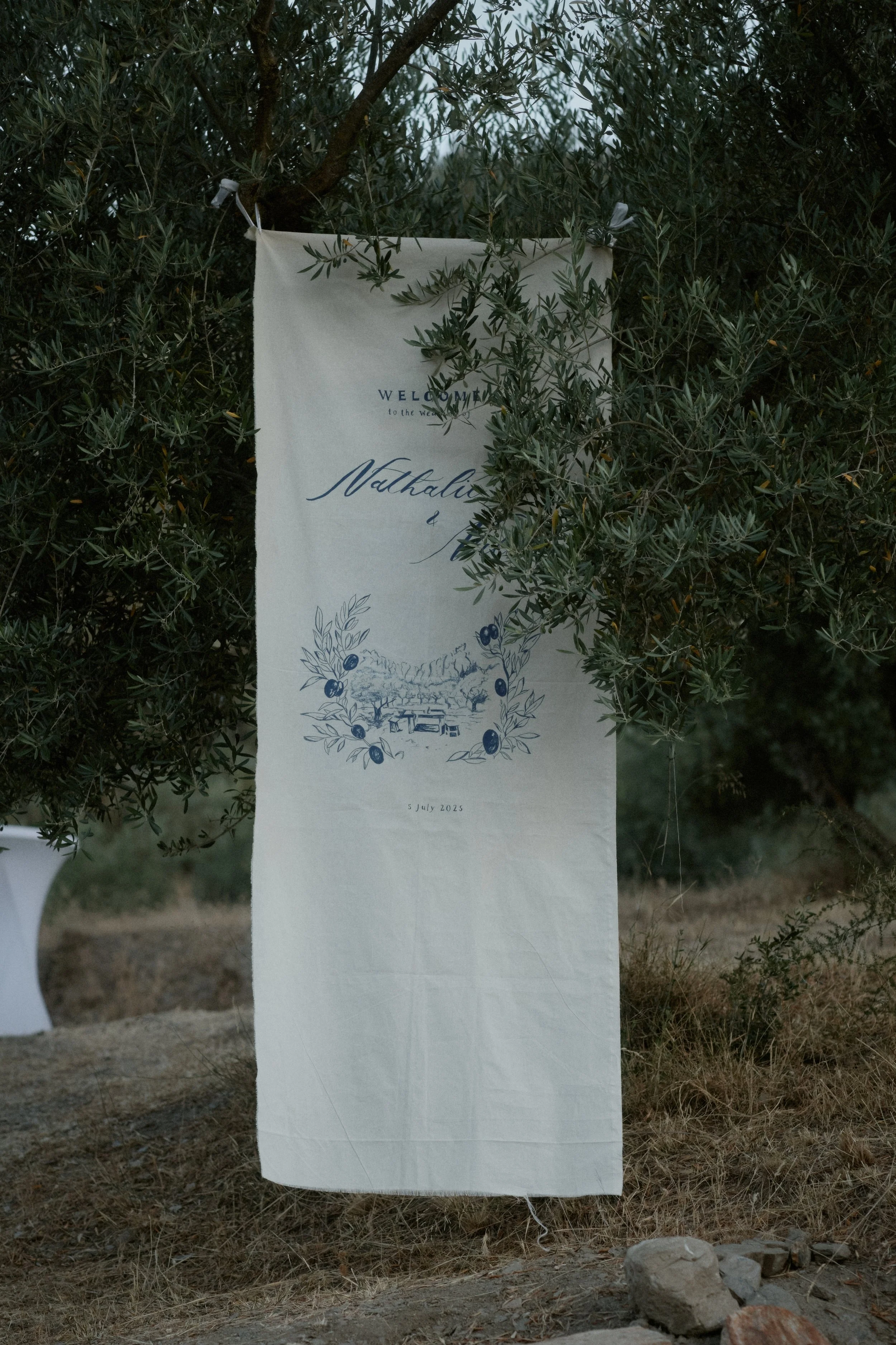 Ein Stoffbanner hängt an einem Baum im Freien, mit blauer Schrift auf weißem Hintergrund, begrüßt die Gäste zu einer Veranstaltung am 5. Juli 2023.