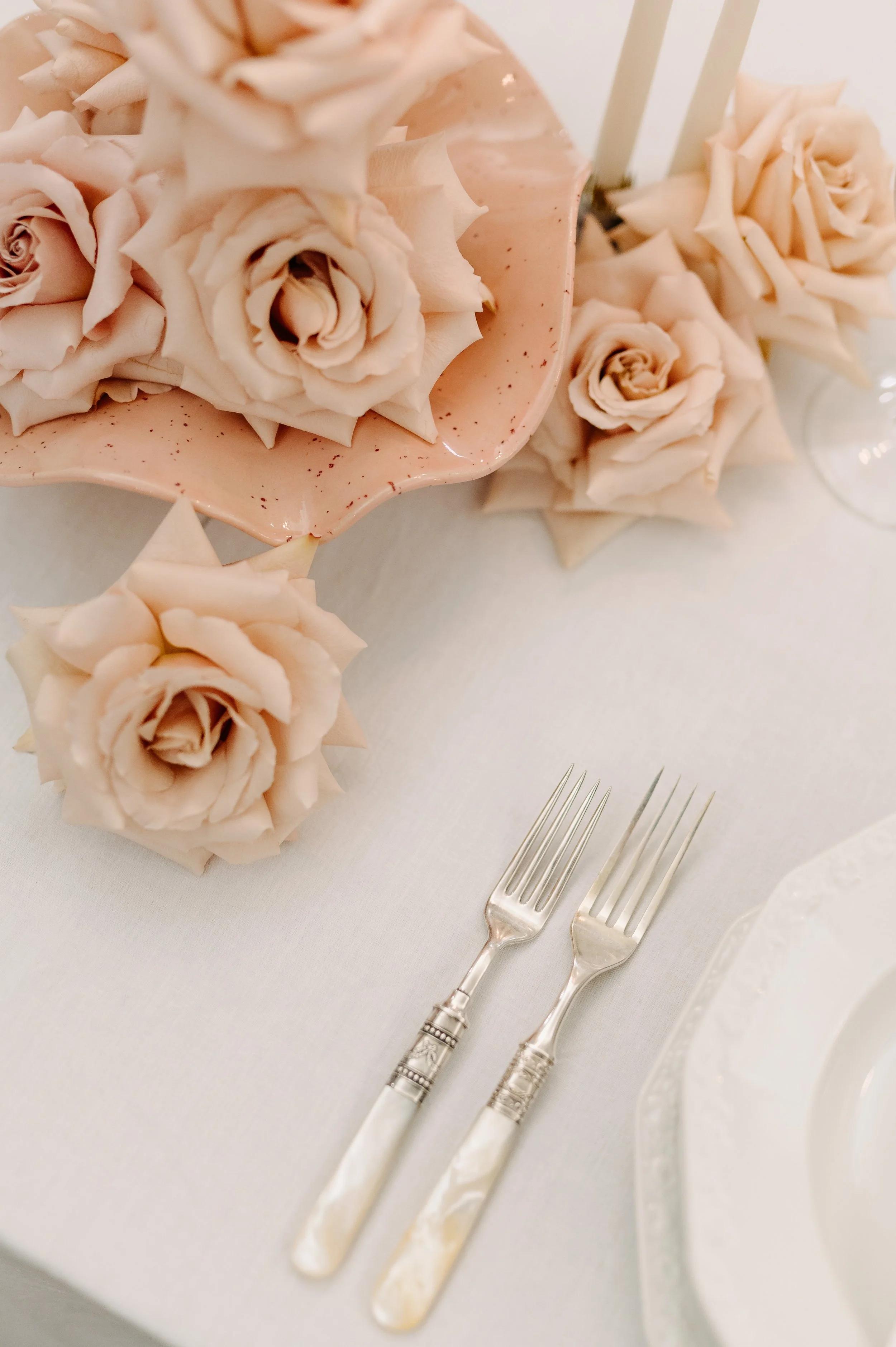 Tischdeko mit hellrosa Rosen, zwei silberne Gabeln, weiße Tischdecke, Spitzentellerrand, eleganter Anlass.