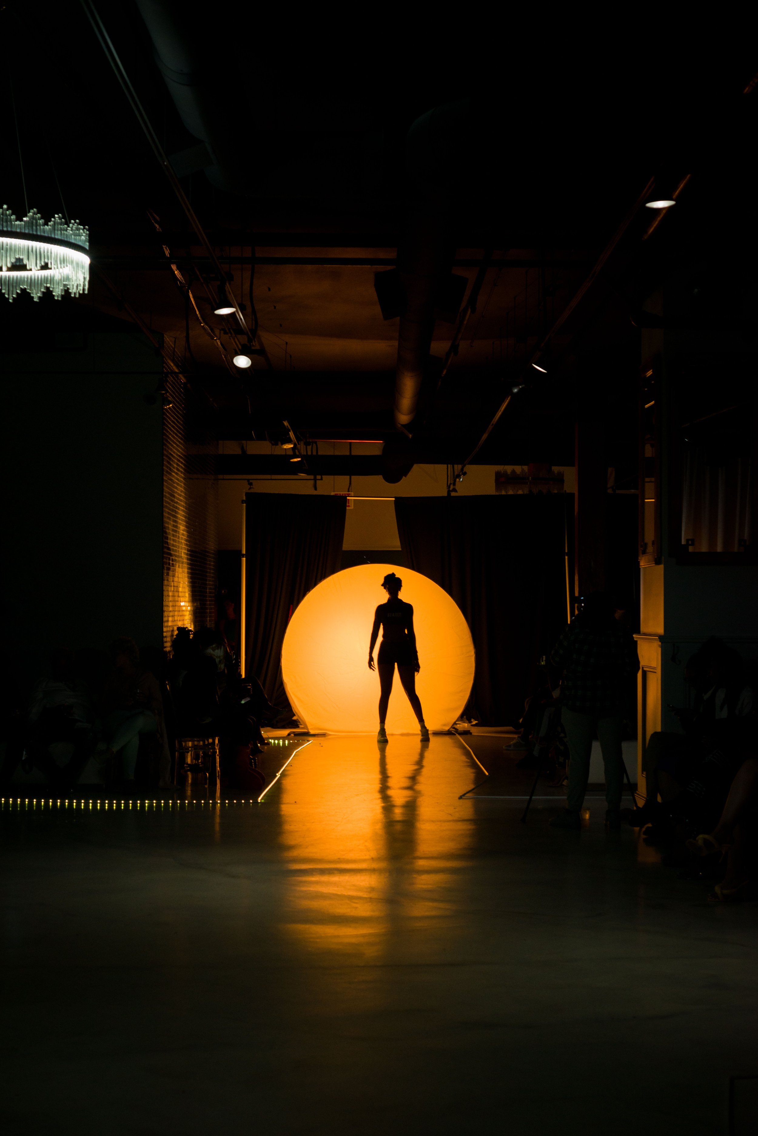 Silhouette of a model on a runway with bright backlighting from a circular light, creating dramatic contrast in a dimly lit room, with an audience on either side.