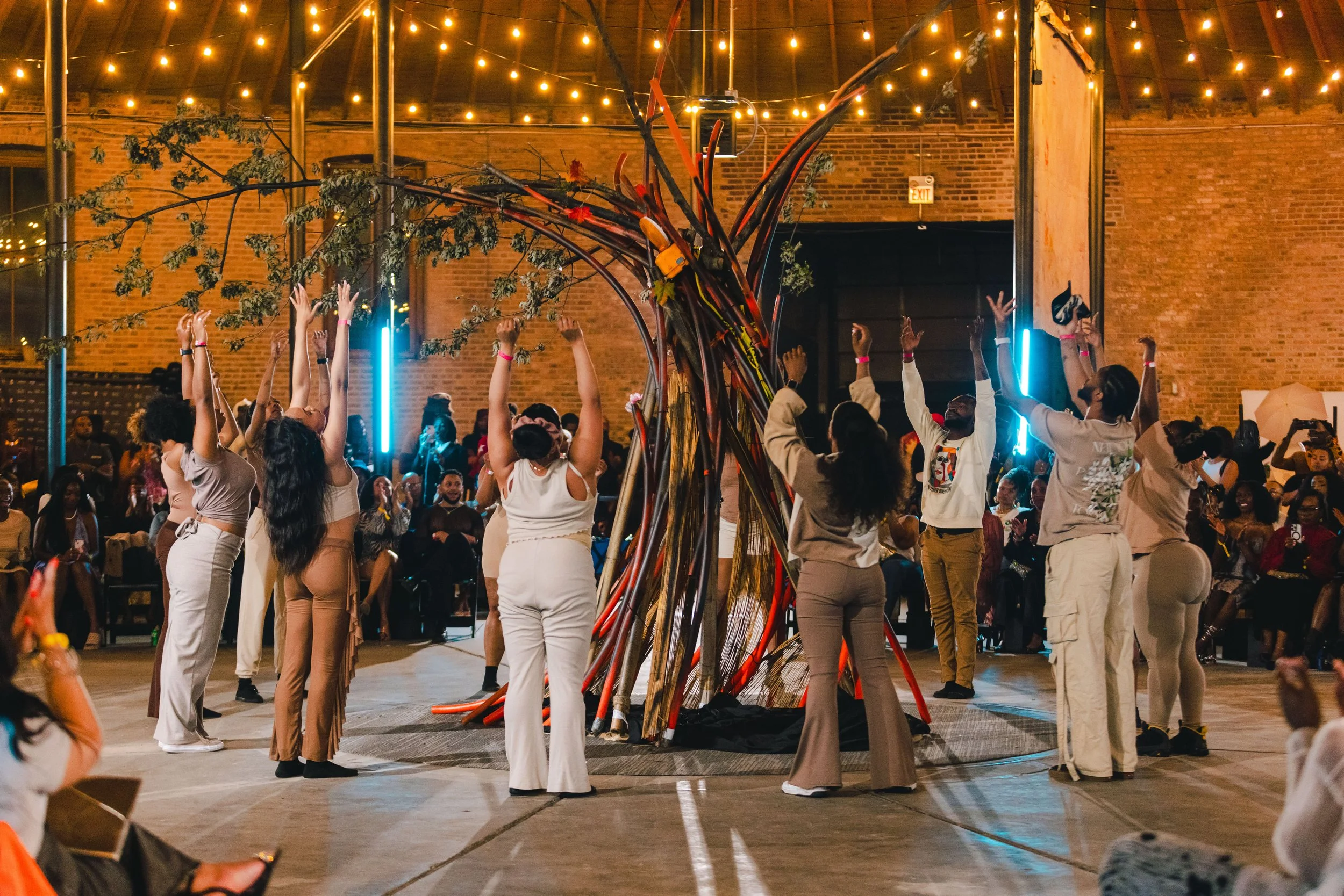 People dancing and celebrating around a colorful tree sculpture inside a venue with string lights and brick walls.