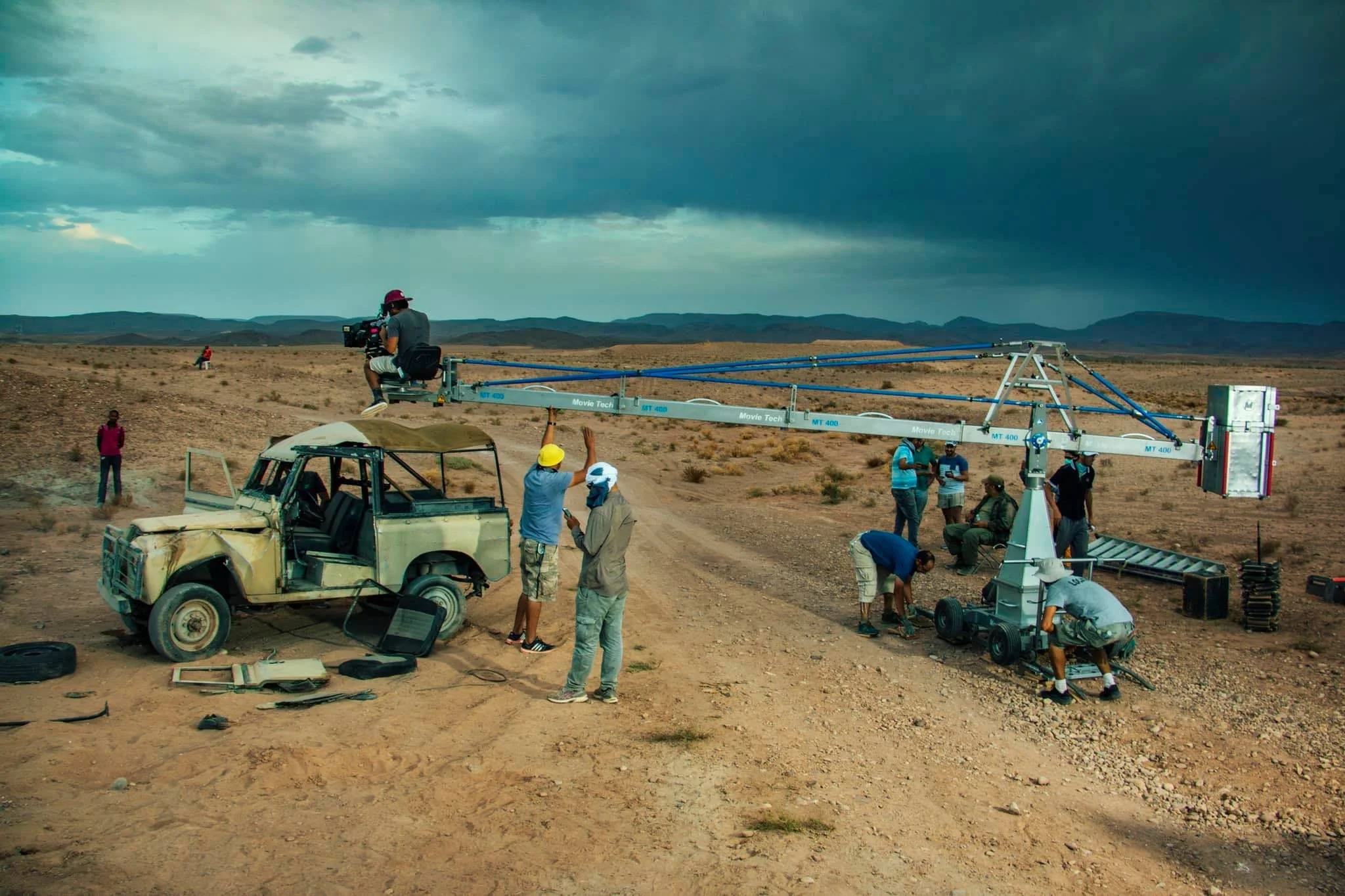 A film crew works on a desert set with a camera mounted on a crane. Crew members adjust equipment around an old, weathered vehicle while others stand near the camera crane. The scene is set in an arid landscape with mountains in the distance and a st