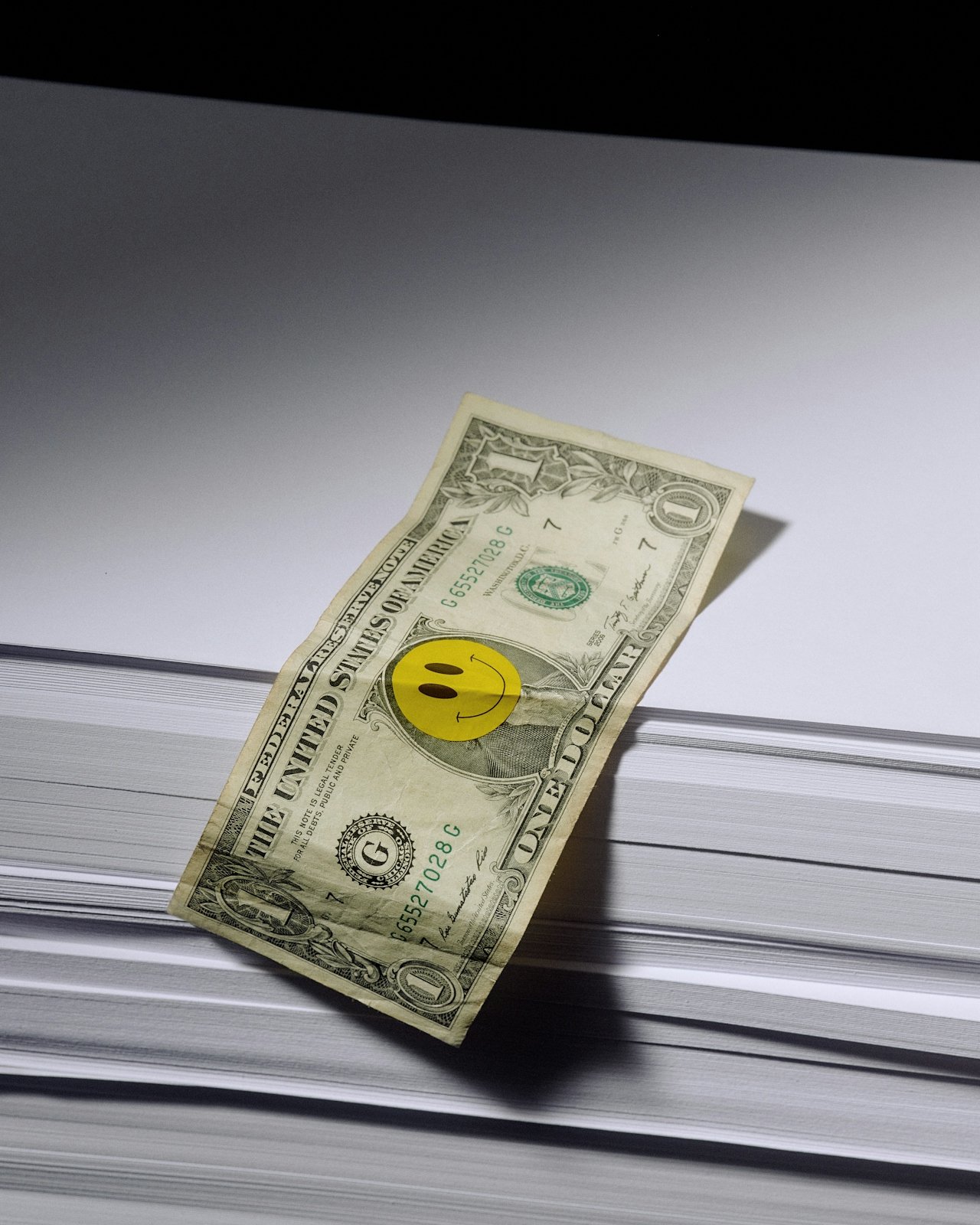 A one-dollar bill with a smiley face sticker on it, resting on a large stack of papers or books.