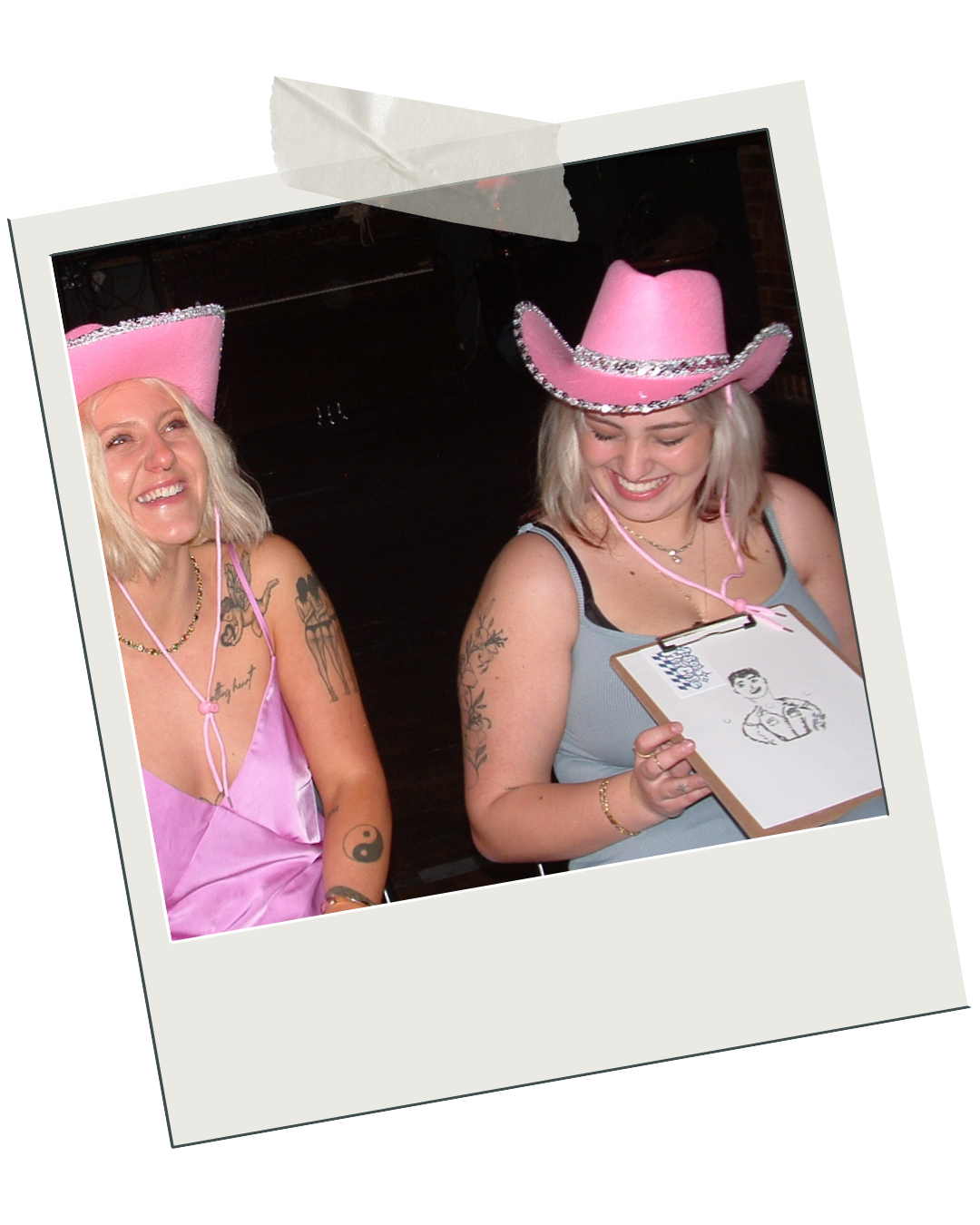 two young blonde women sitting next to eachother at a hens party, they are both laughing and wearing pink cowgirl hats. the one on the right is holding a clipboard with a drawing of a police officer