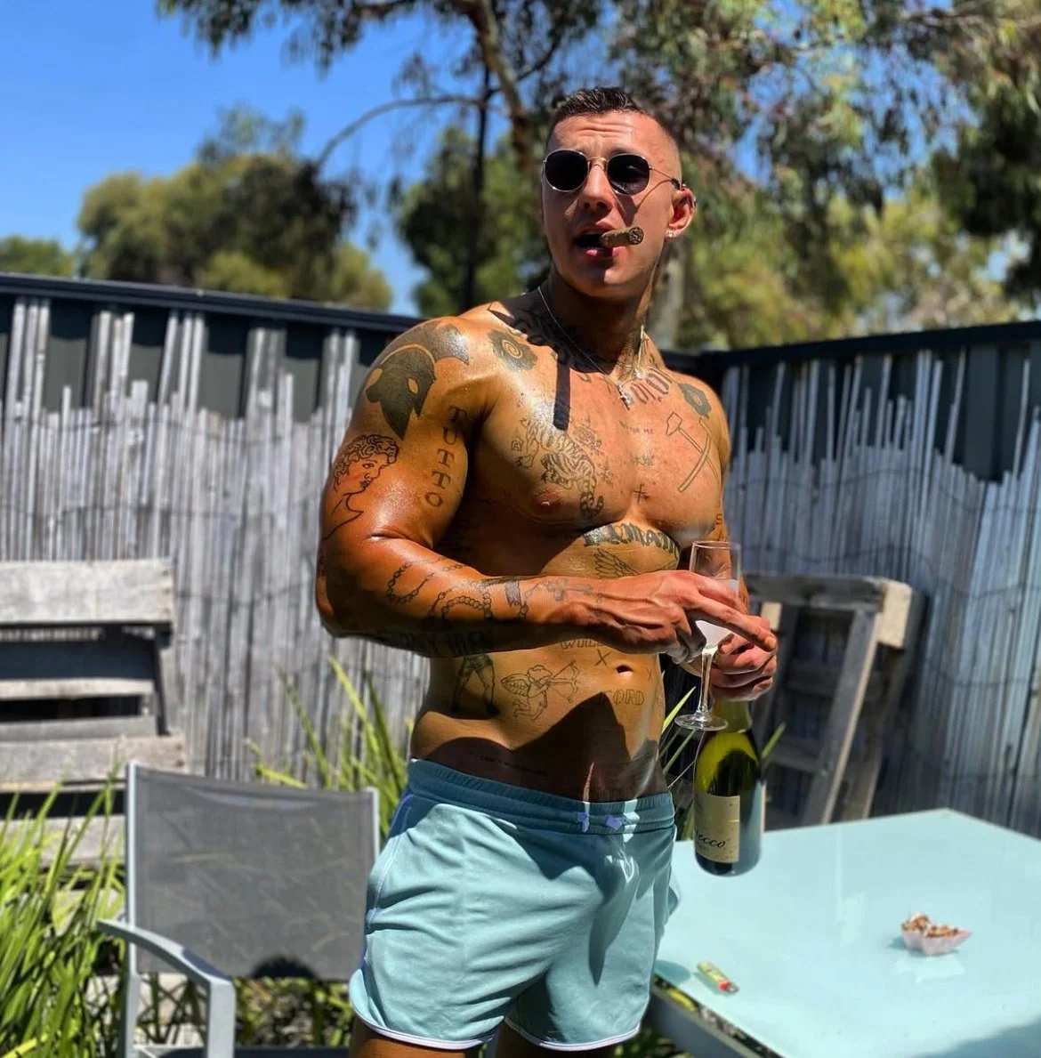 Dirk Diggler posing in beach shorts at a pool party holding a flute and bottle of champagne -male entertainer for Melbourne hens parties and girls’ nights.