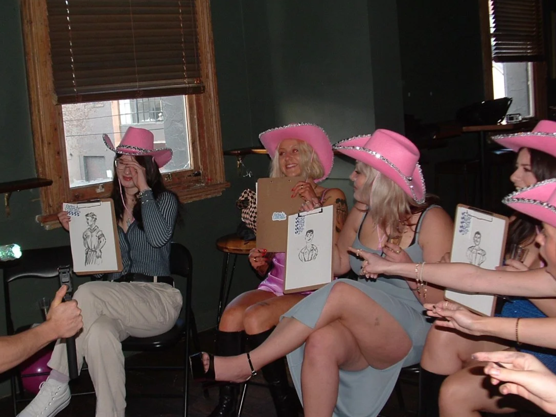 a group of young women sitting in side by side in a green room wearing pink cowgirl hats and holding up clipboards with drawings of a male model, many of the girls are pointing to the drawing in the far left side of the picture