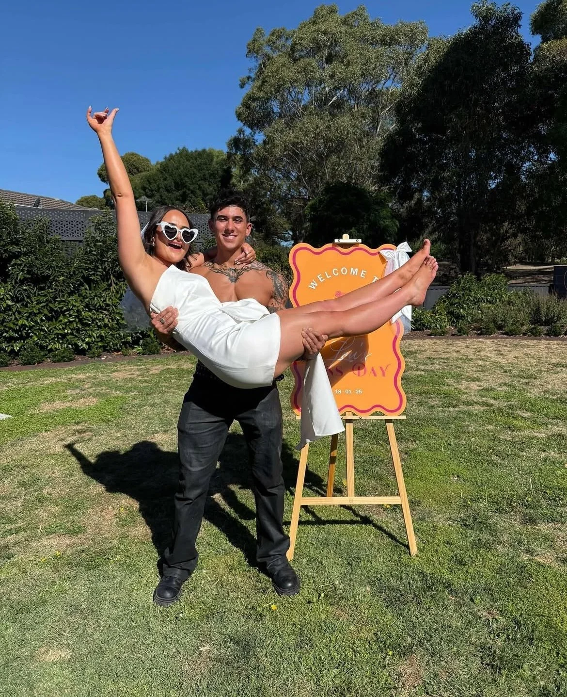 A man lifting a woman in a white dress, both smiling, outdoors near a sign with a pink and orange design, trees, and a clear blue sky.