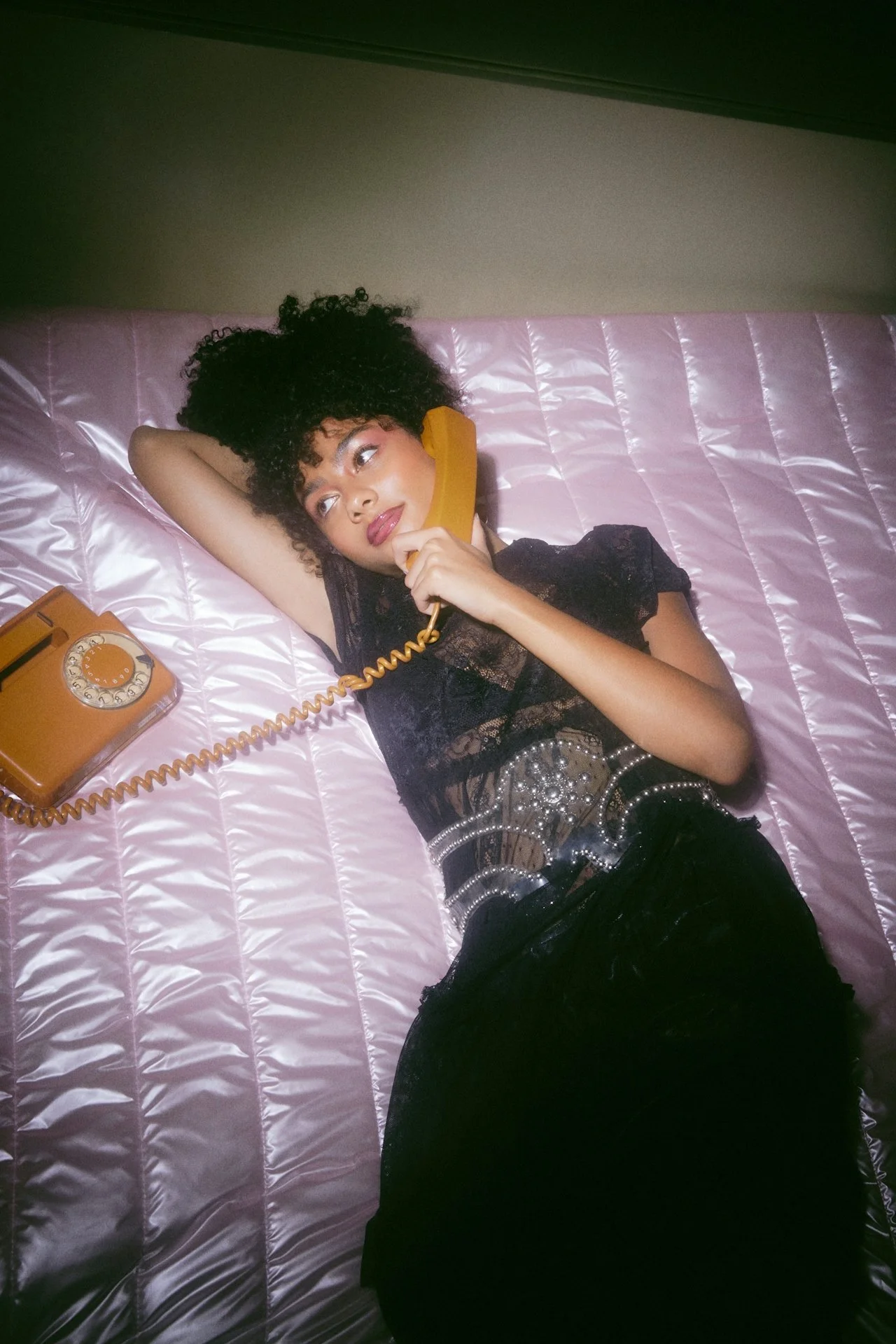 A woman with curly hair lying on a pink quilted bed, talking on a yellow rotary phone, wearing a black lace top and skirt with a pearl belt.
