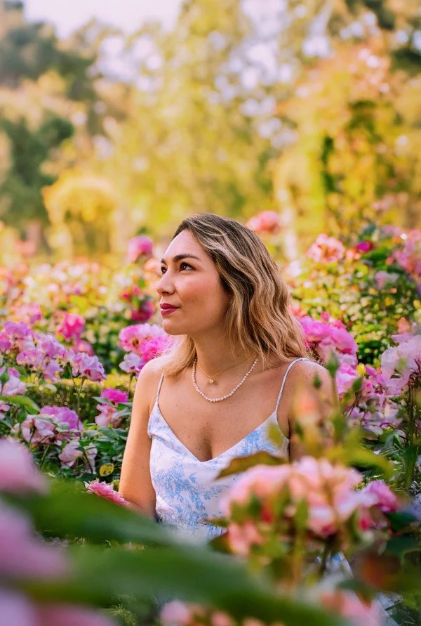 A woman with wavy hair wearing a light blue floral dress and a pearl necklace, sitting among pink and purple flowers in a garden on a sunny day.