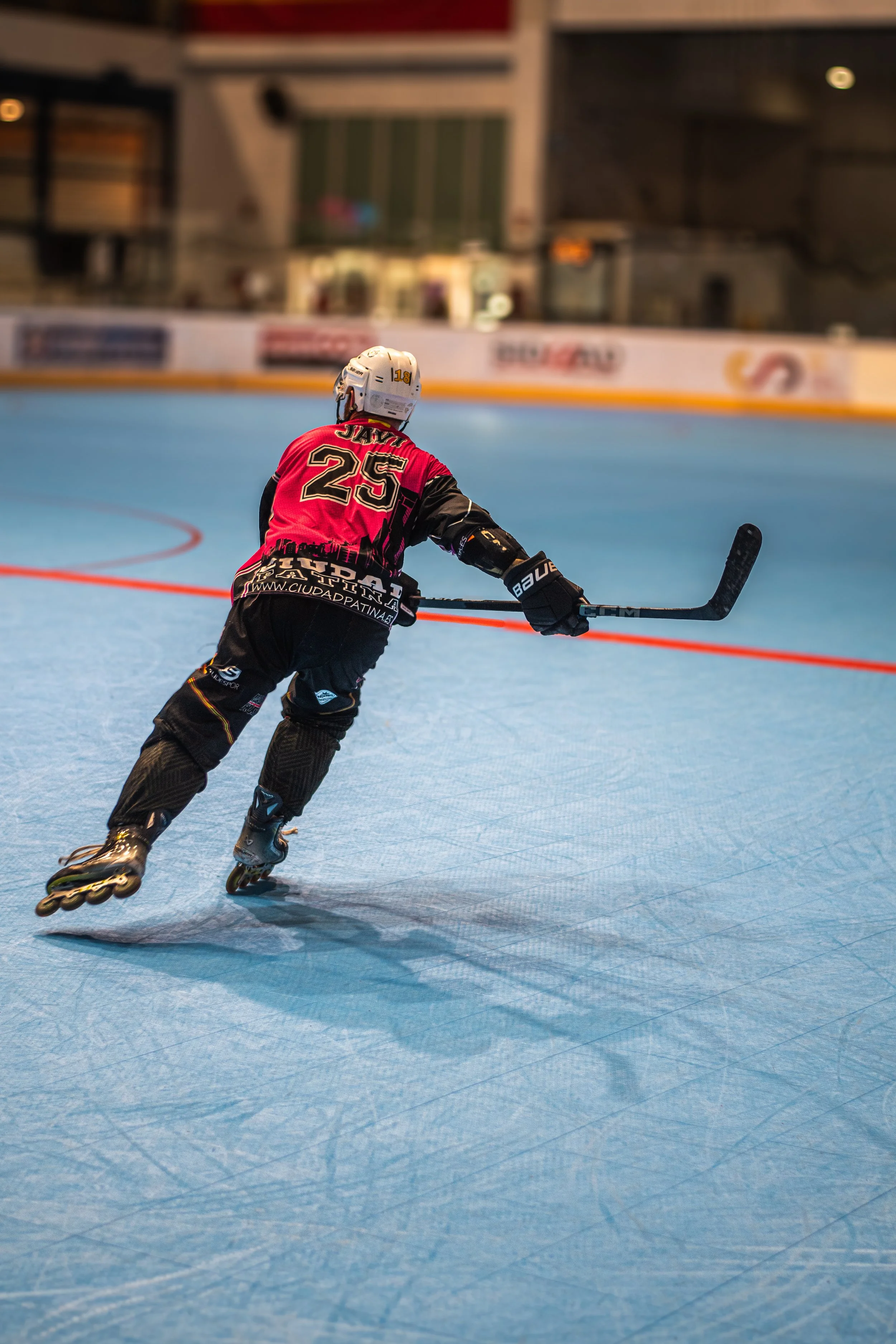 A roller hockey player dressed in black and red gear, wearing a helmet, skating on a blue rink surface, holding a hockey stick.