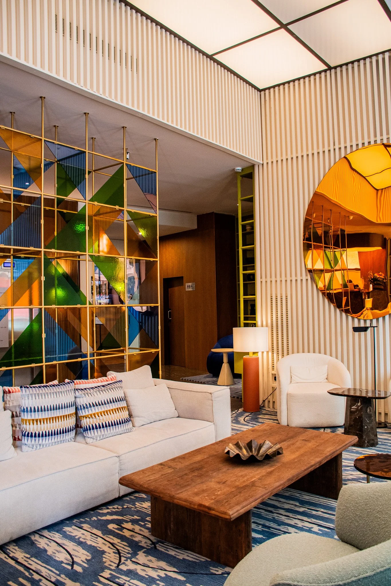 A modern hotel lobby with a white sofa, colorful pillows, a wooden coffee table, a round mirror on a textured wall, and a decorative room divider with geometric patterns.
