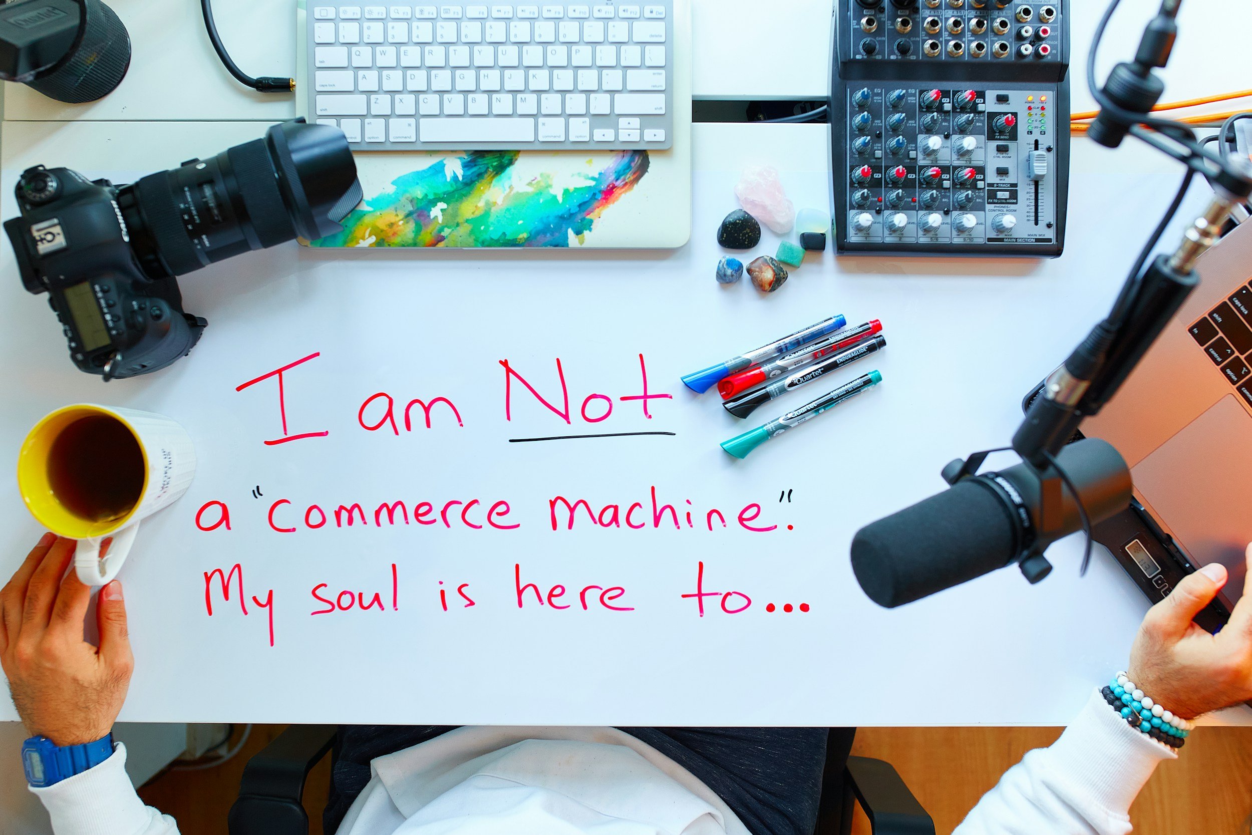 A white desk with cameras, a keyboard, a mixing console, colorful markers, a coffee mug, rocks, a laptop, and a handwritten message in red marker that says, "I am Not a 'commerce machine.' My soul is here to..."